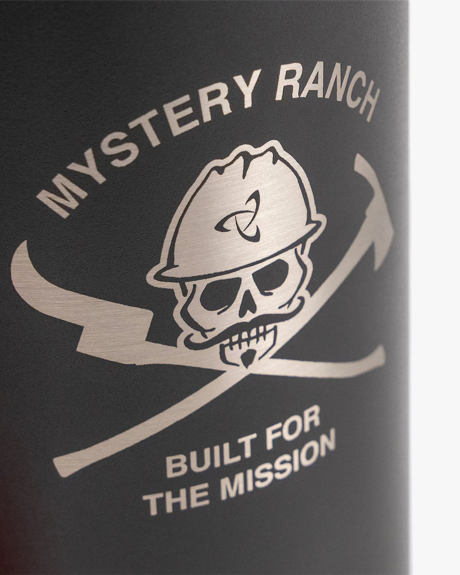 Close-up of a black surface with a silver skull wearing a hard hat and crossed tools, labeled "Mystery Ranch" and "Built for the mission.
