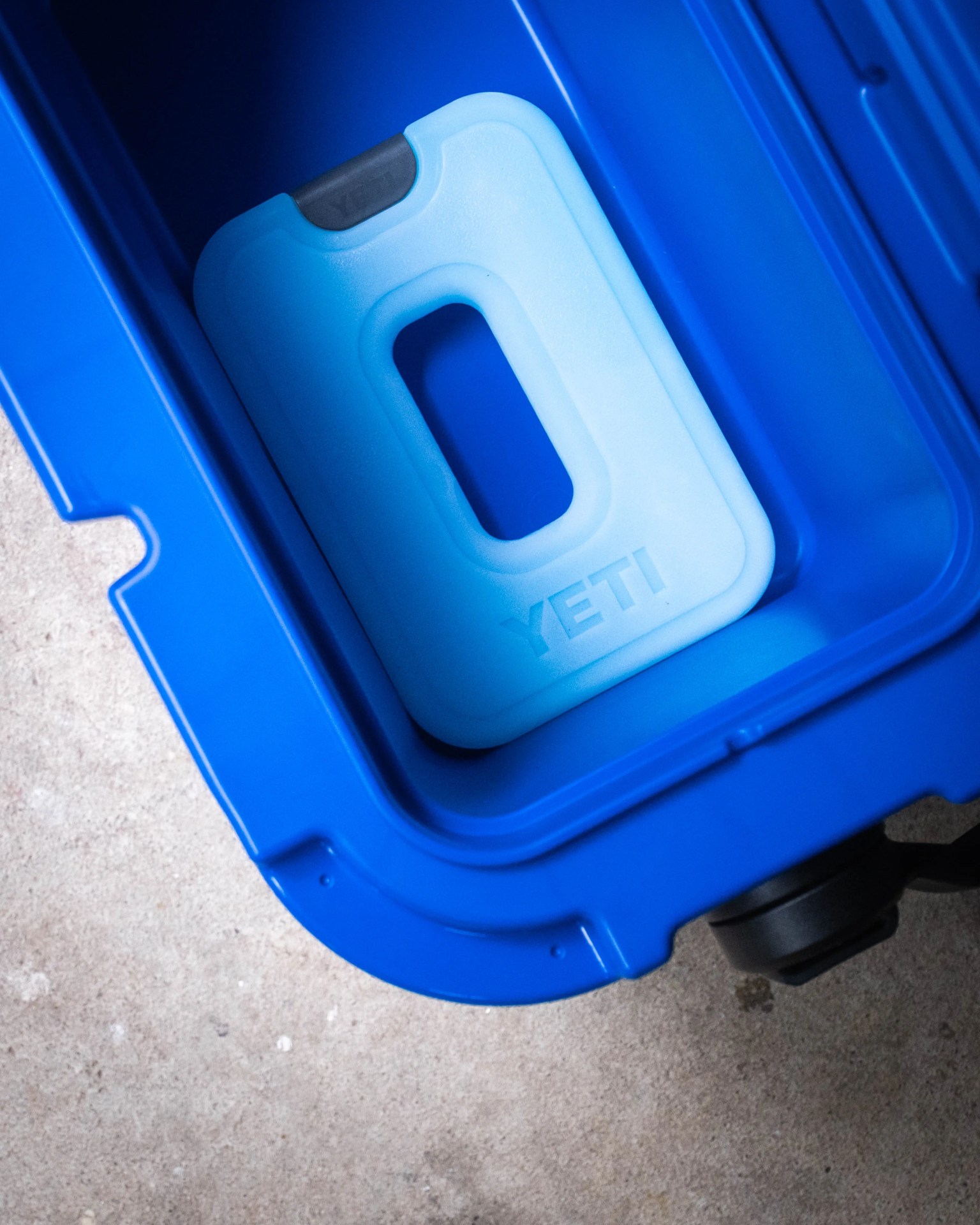 Light blue YETI ice pack inside a bright blue cooler on a concrete floor.