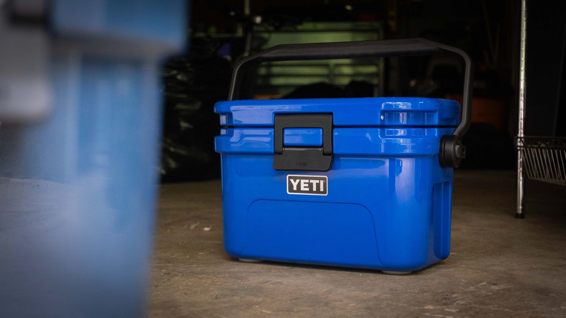 Blue YETI cooler with black handle and latch on a concrete floor in a dimly lit room.