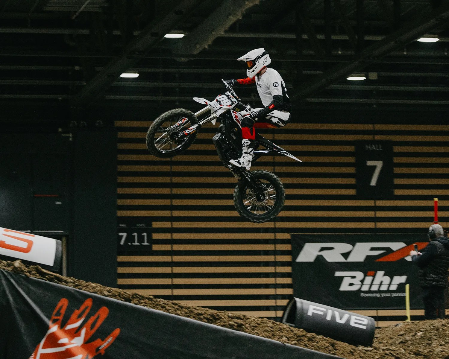 Motocross rider in white gear performing a jump on a dirt track inside an indoor arena.