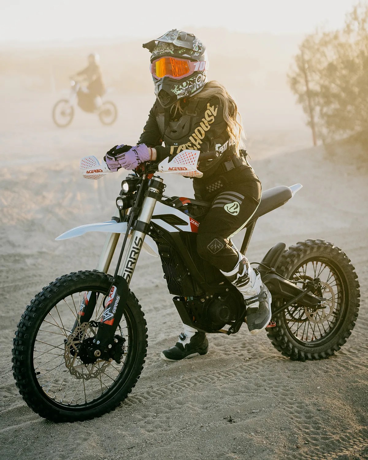 Person wearing black motocross gear and orange goggles sitting on a black and white electric dirt bike on sandy terrain.