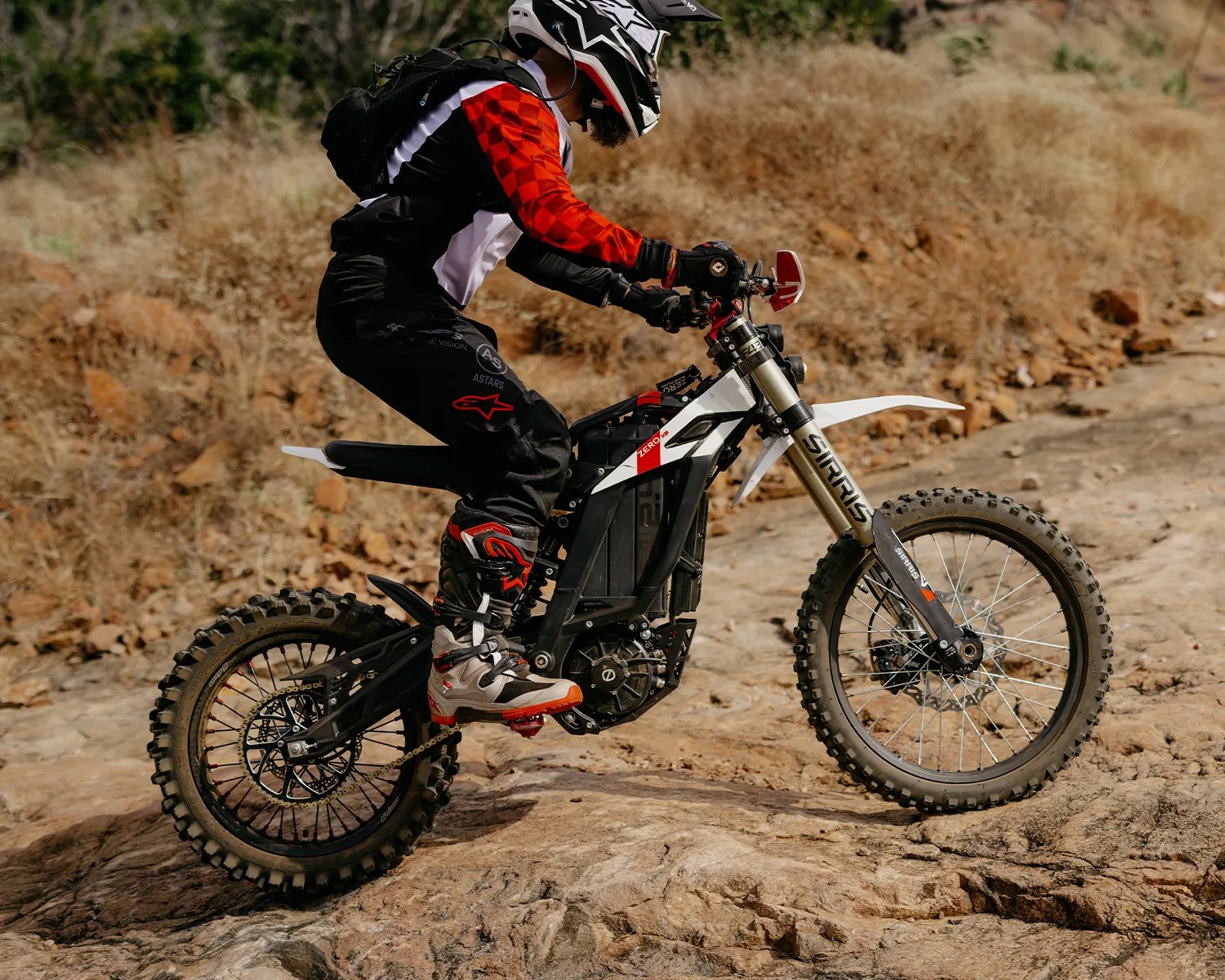 Person wearing a red and black motocross outfit riding a white and black electric dirt bike on rocky terrain.
