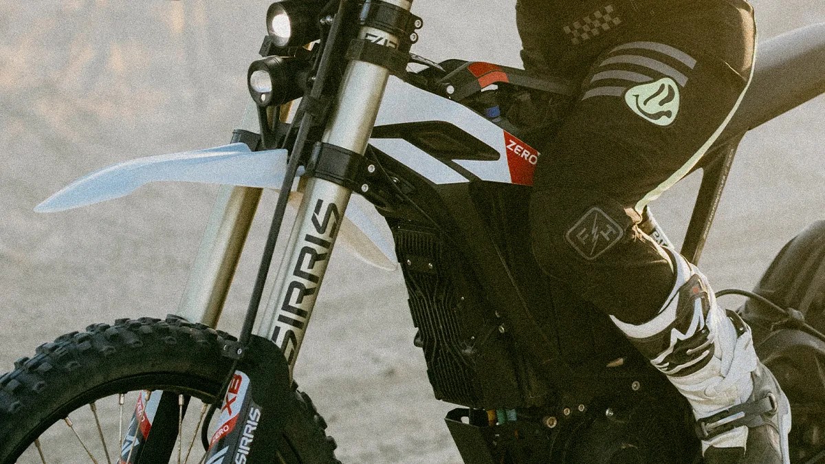 Close-up of a black and white Sur-Ron electric dirt bike with knobby tires and a rider wearing black pants and white motocross boots.