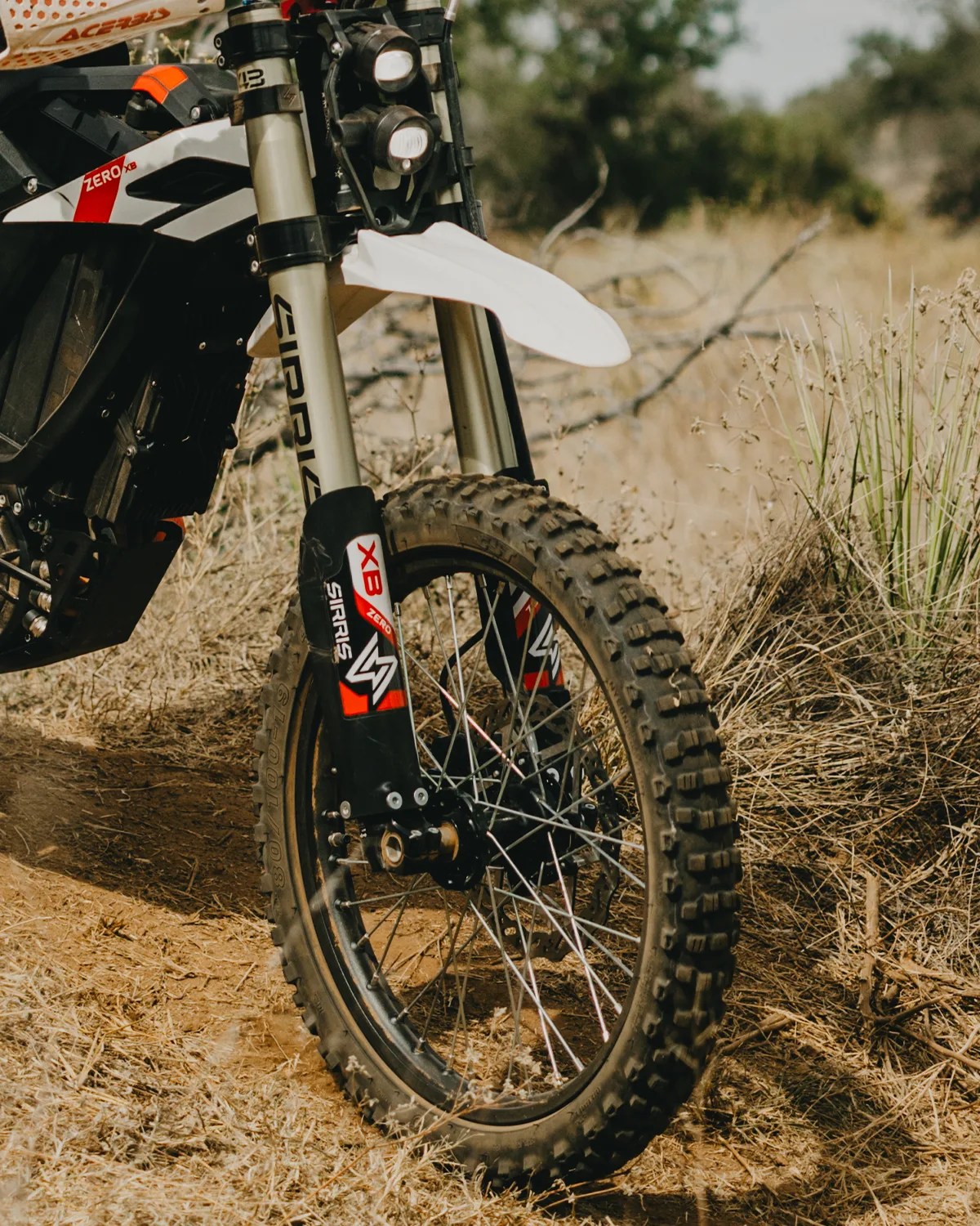 Front wheel and suspension fork of a dirt bike with knobby tire on a dry, grassy trail.
