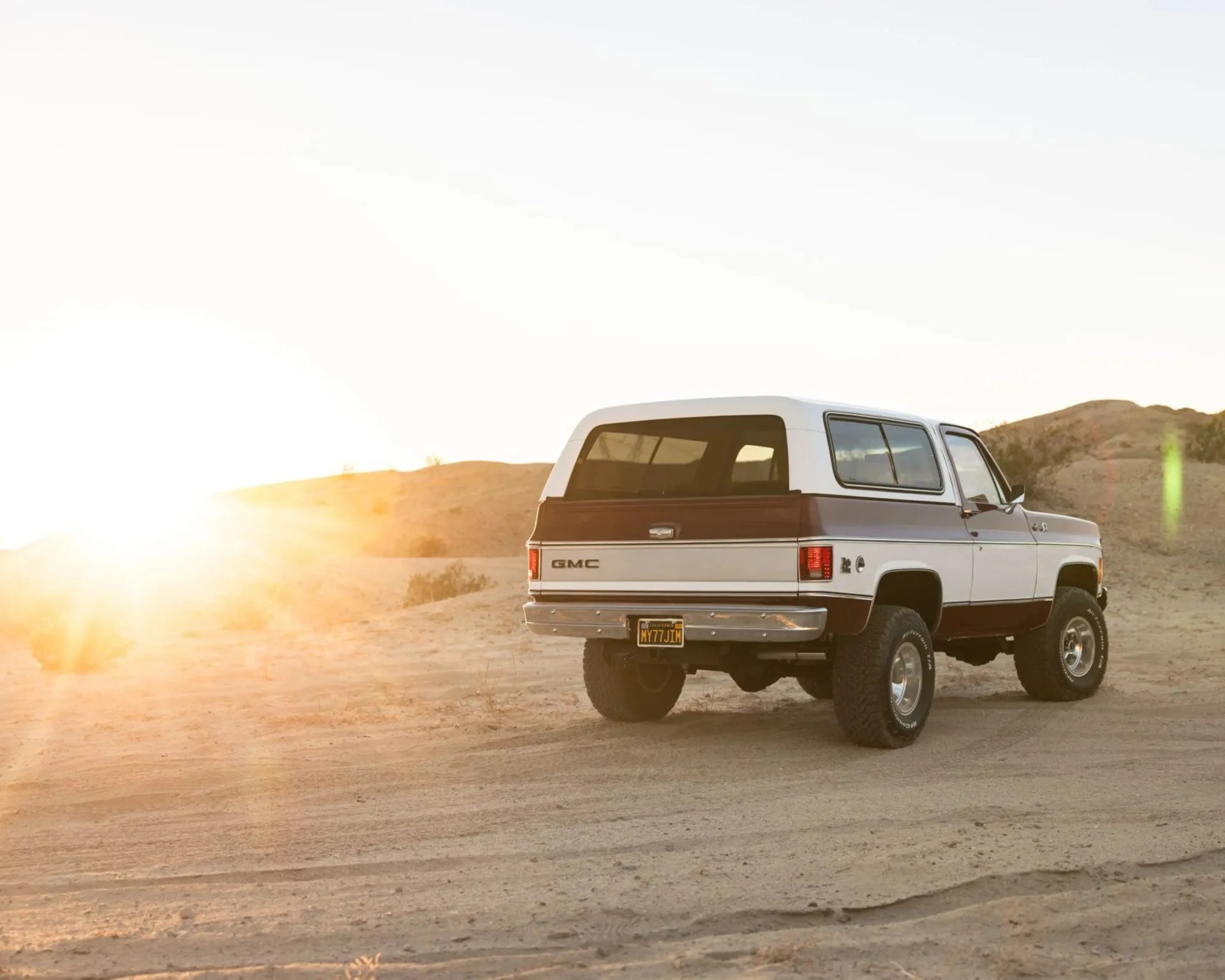 White and brown GMC SUV with tinted windows parked on a dirt road at sunset in a desert landscape.