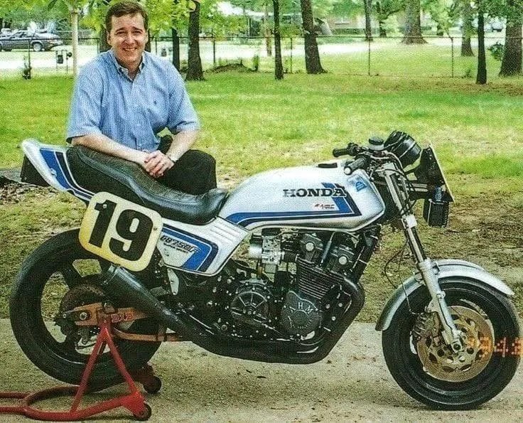 Man in a blue shirt sitting on a white and blue Honda racing motorcycle with number 19 on the side.