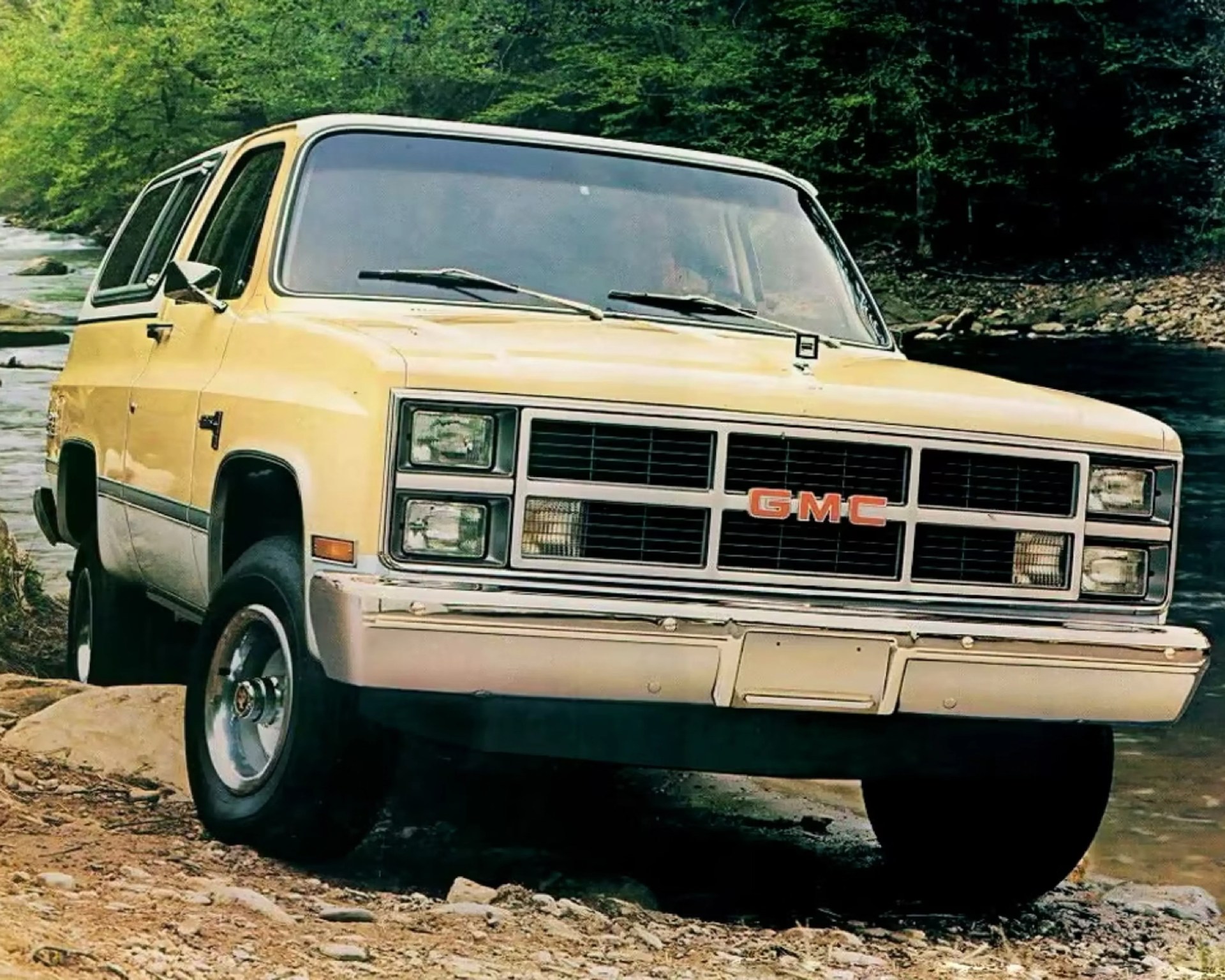 Yellow and beige GMC SUV parked on rocky terrain near a river with green trees in the background.