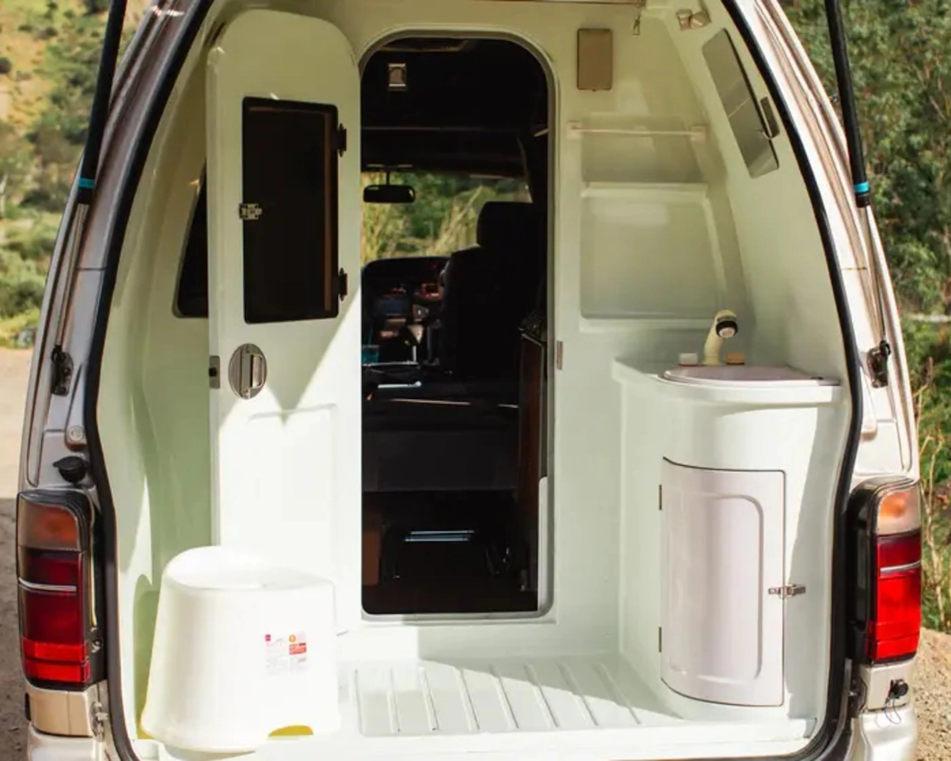 White portable camping toilet and bucket inside the rear of a van with open door.