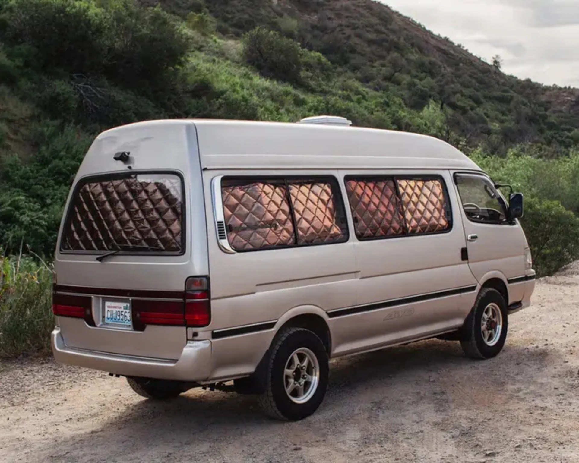 White camper van with quilted window covers parked on a dirt road near green hills.