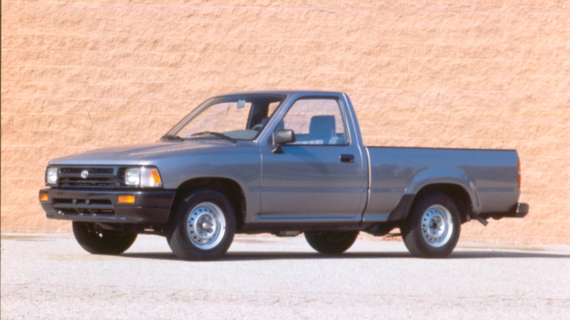 Blue single-cab pickup truck parked on a light gravel surface with a beige brick wall background.
