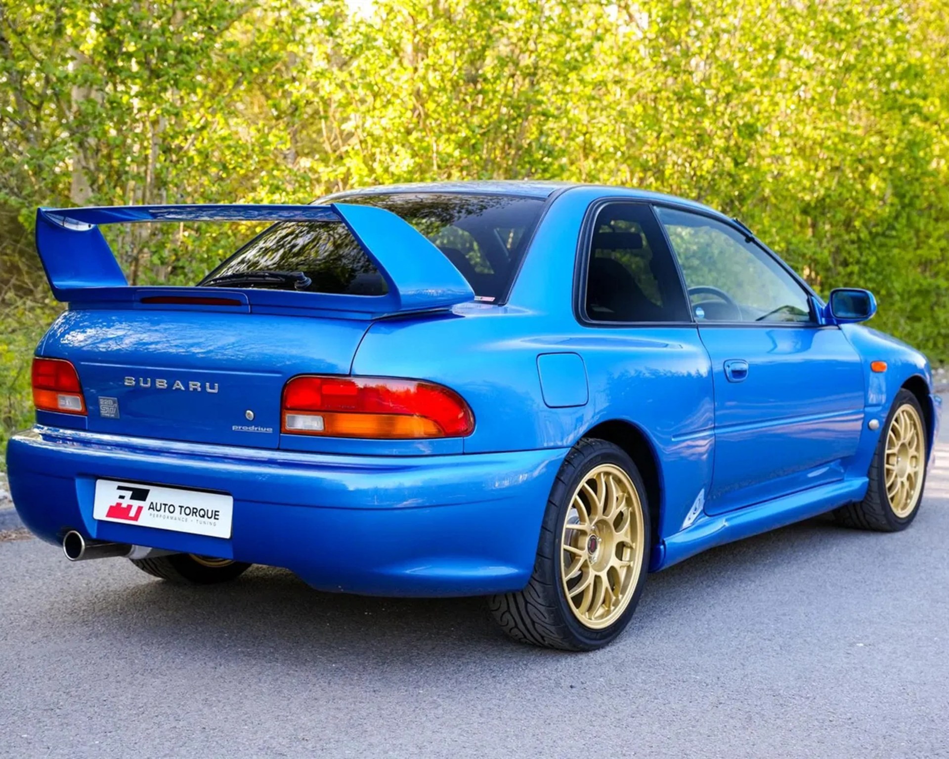 Blue Subaru sports car with large rear spoiler and gold alloy wheels parked on a road.