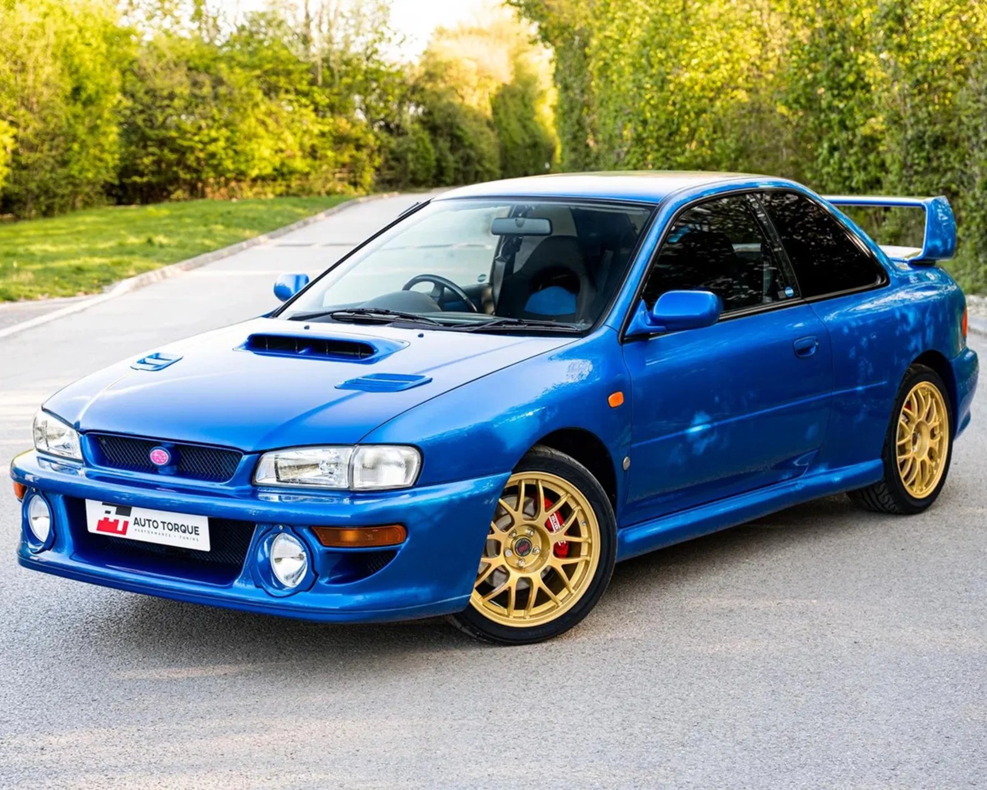 Blue Subaru Impreza WRX with gold alloy wheels parked on a road with green trees in the background.