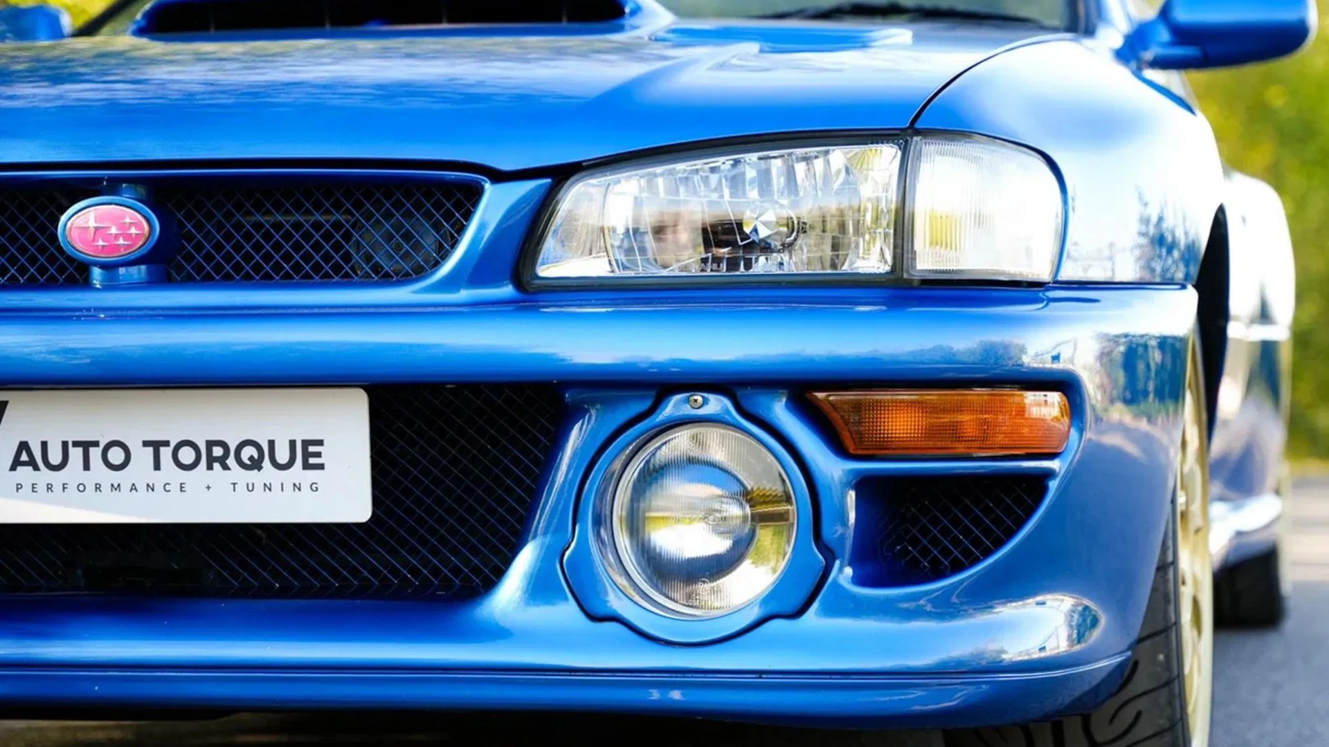 Front view of a blue Subaru car showing the grille with Subaru emblem, headlight, fog light, and a license plate reading "AUTO TORQUE PERFORMANCE + TUNING.