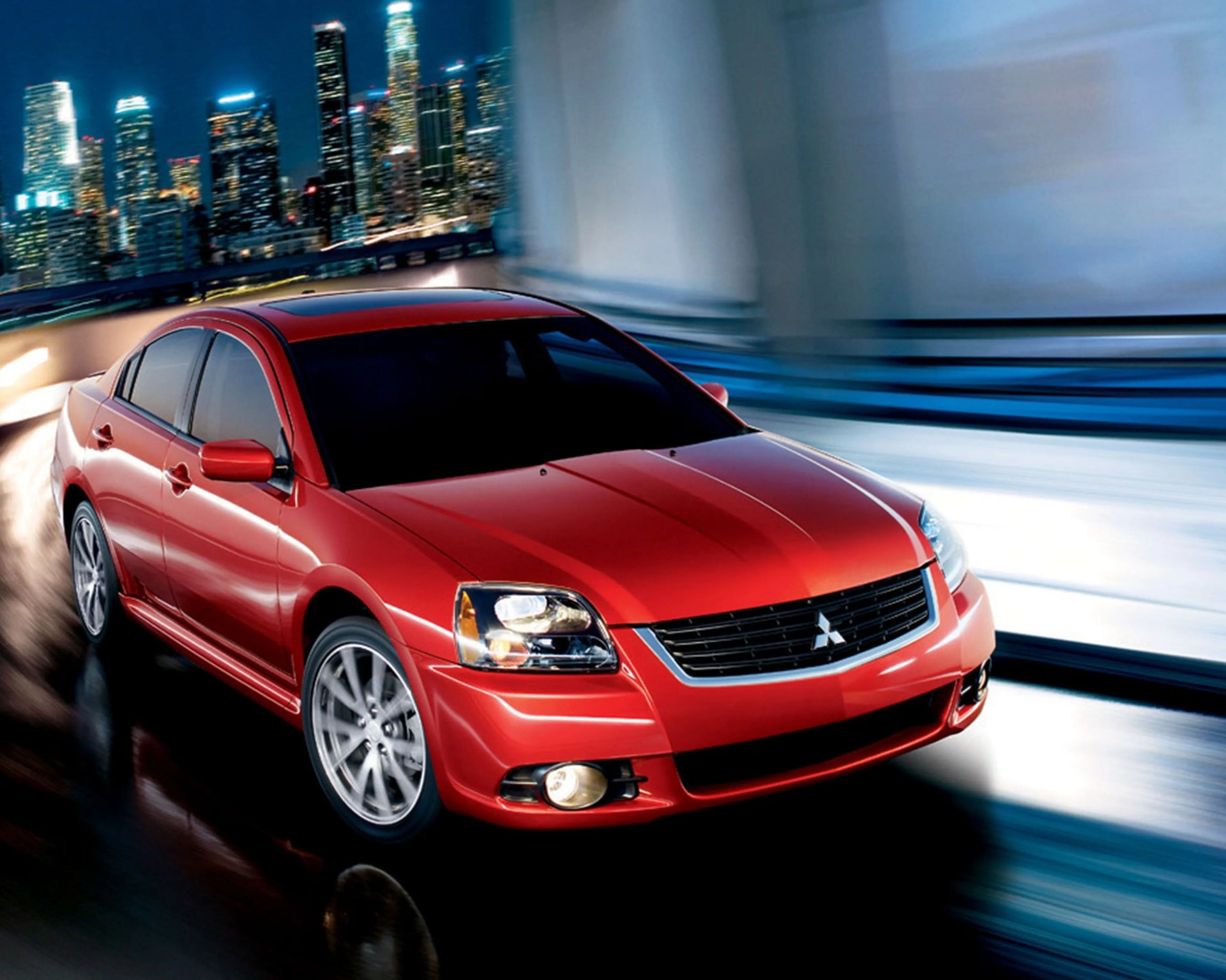 Red Mitsubishi sedan with sunroof driving on a curved road at night with city skyline in the background.