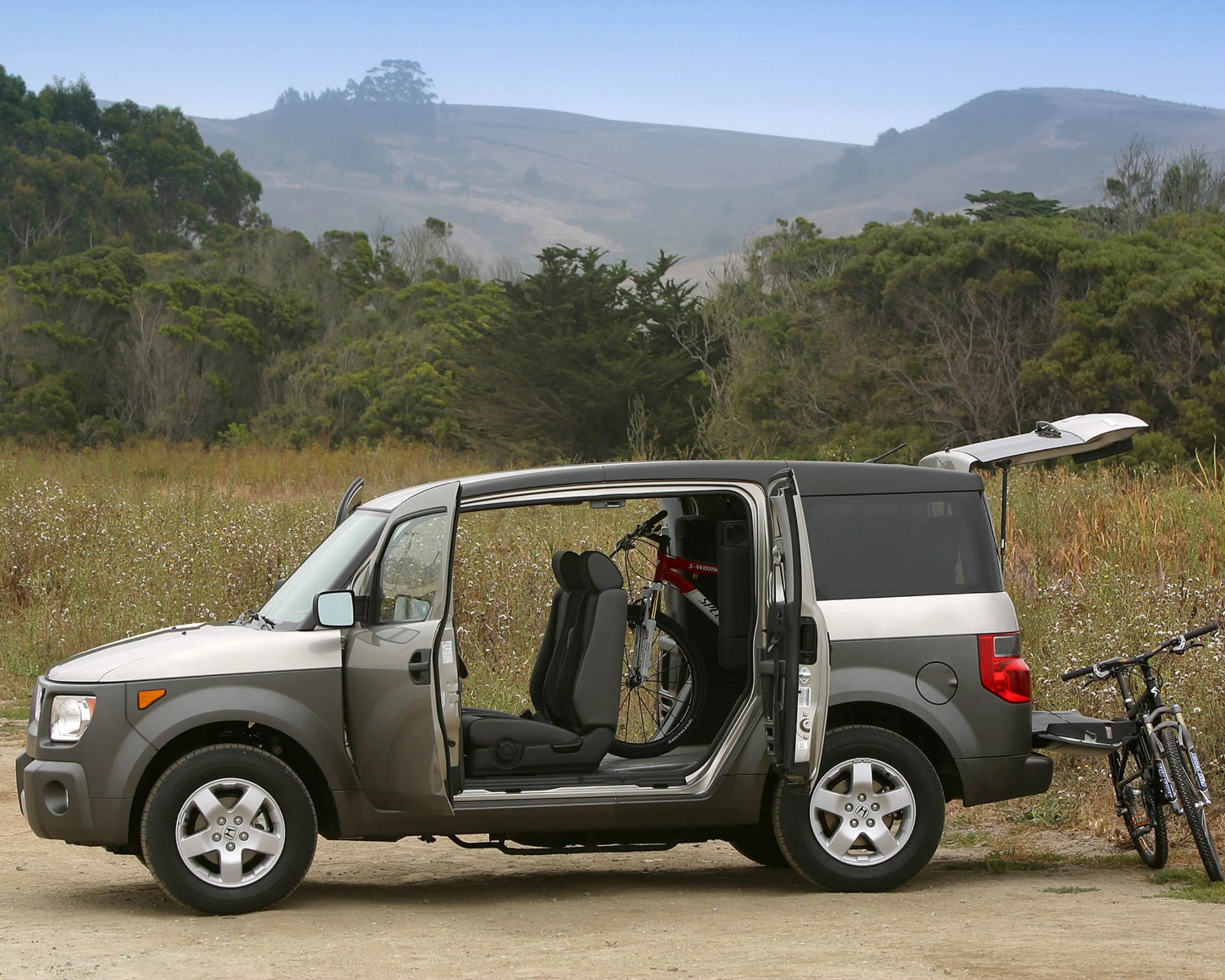 Silver and black Honda Element with open doors and hatchback, carrying a red bicycle inside and a black bicycle on a rear rack.