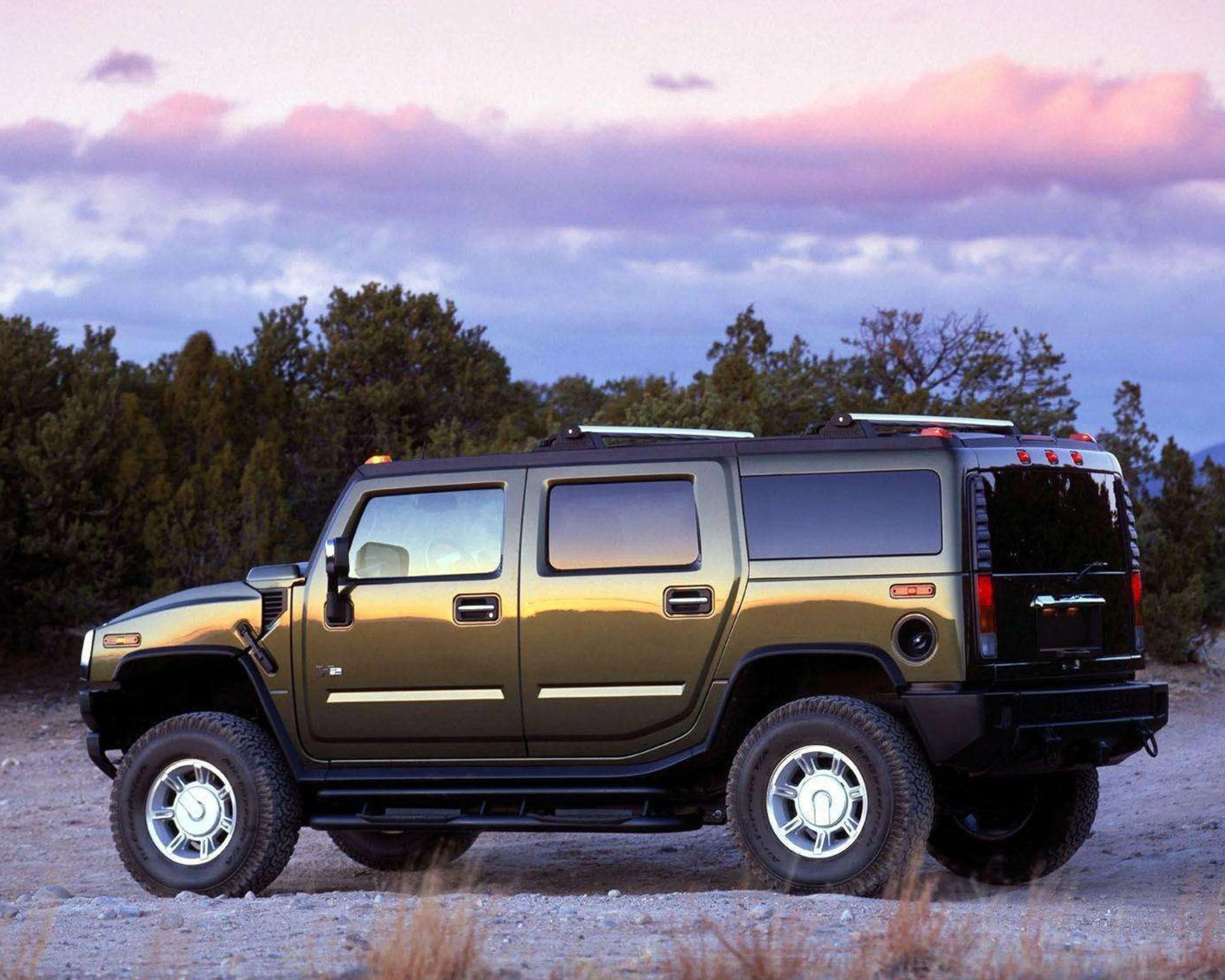 Olive green Hummer H2 SUV parked on rocky terrain with trees and a pink-purple sky in the background.