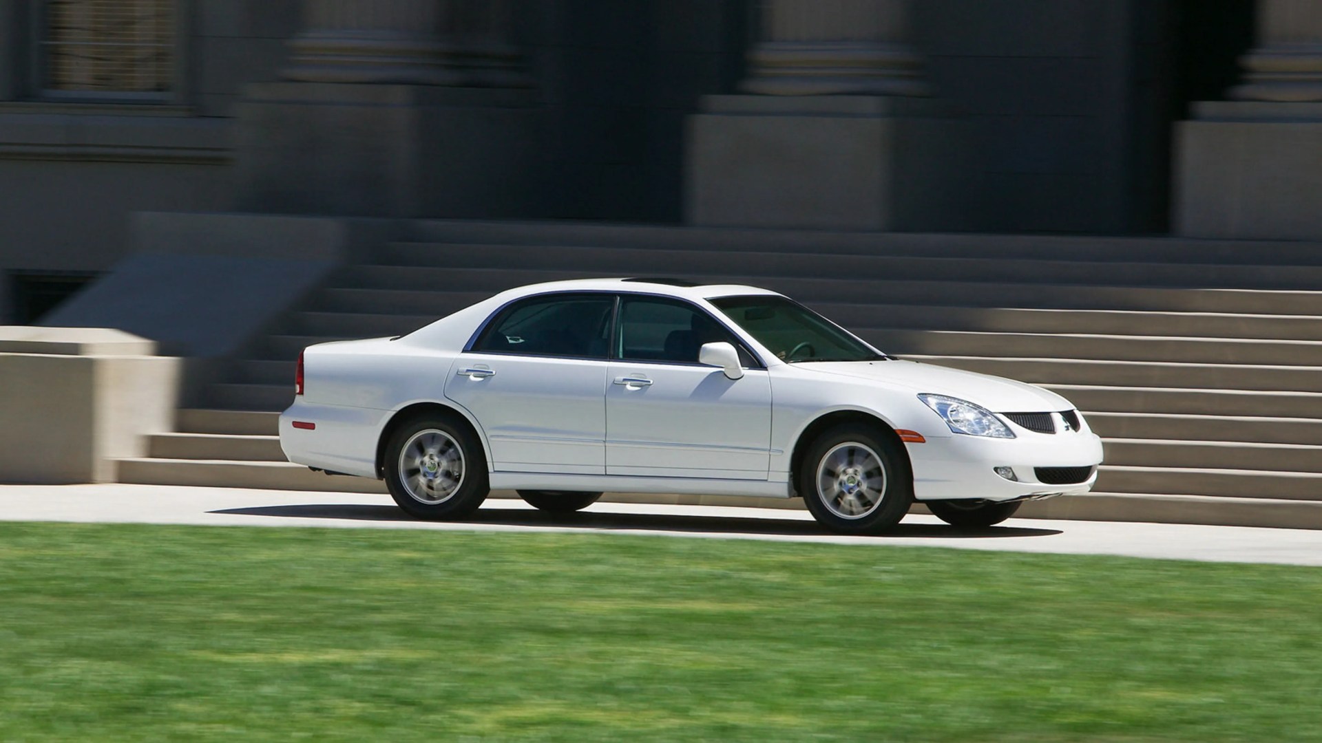 White four-door sedan driving on a paved path with stone steps and columns in the background.