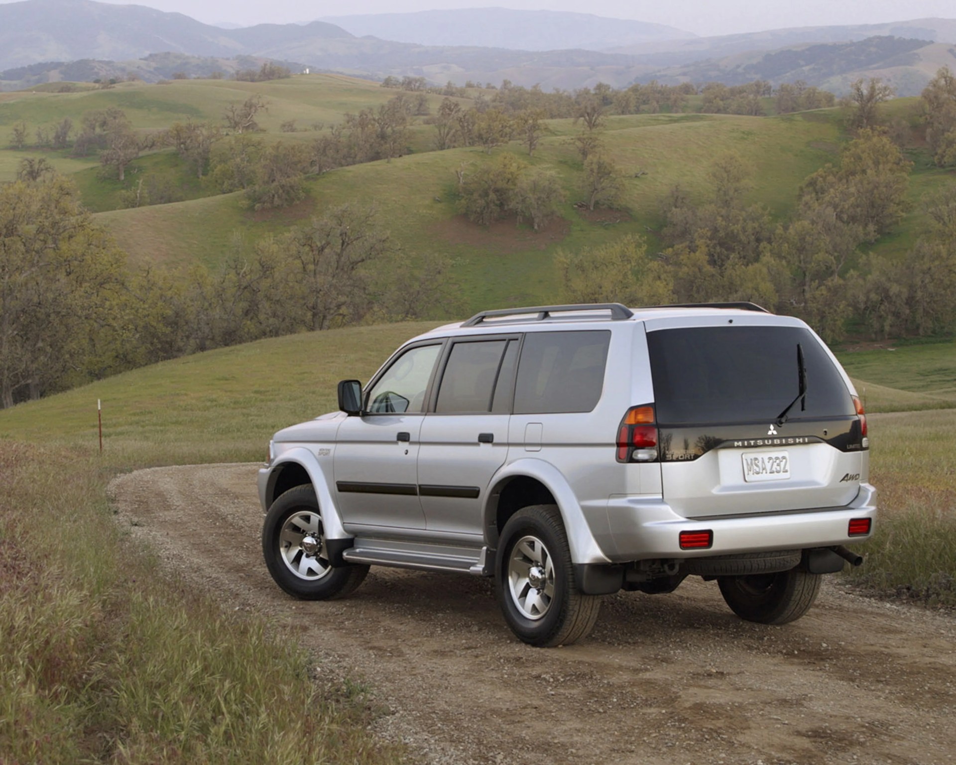 Silver Mitsubishi SUV parked on a dirt road with green hills and trees in the background.