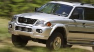 Silver Mitsubishi SUV with roof rack driving on a dirt road with blurred greenery background.