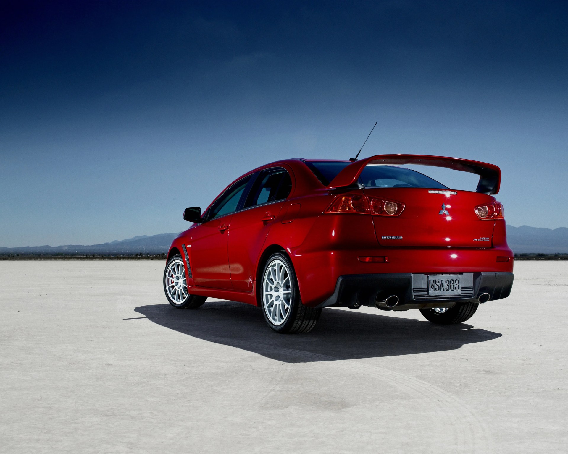 Red Mitsubishi Lancer Evolution with large rear spoiler parked on a flat, open surface under a clear blue sky.