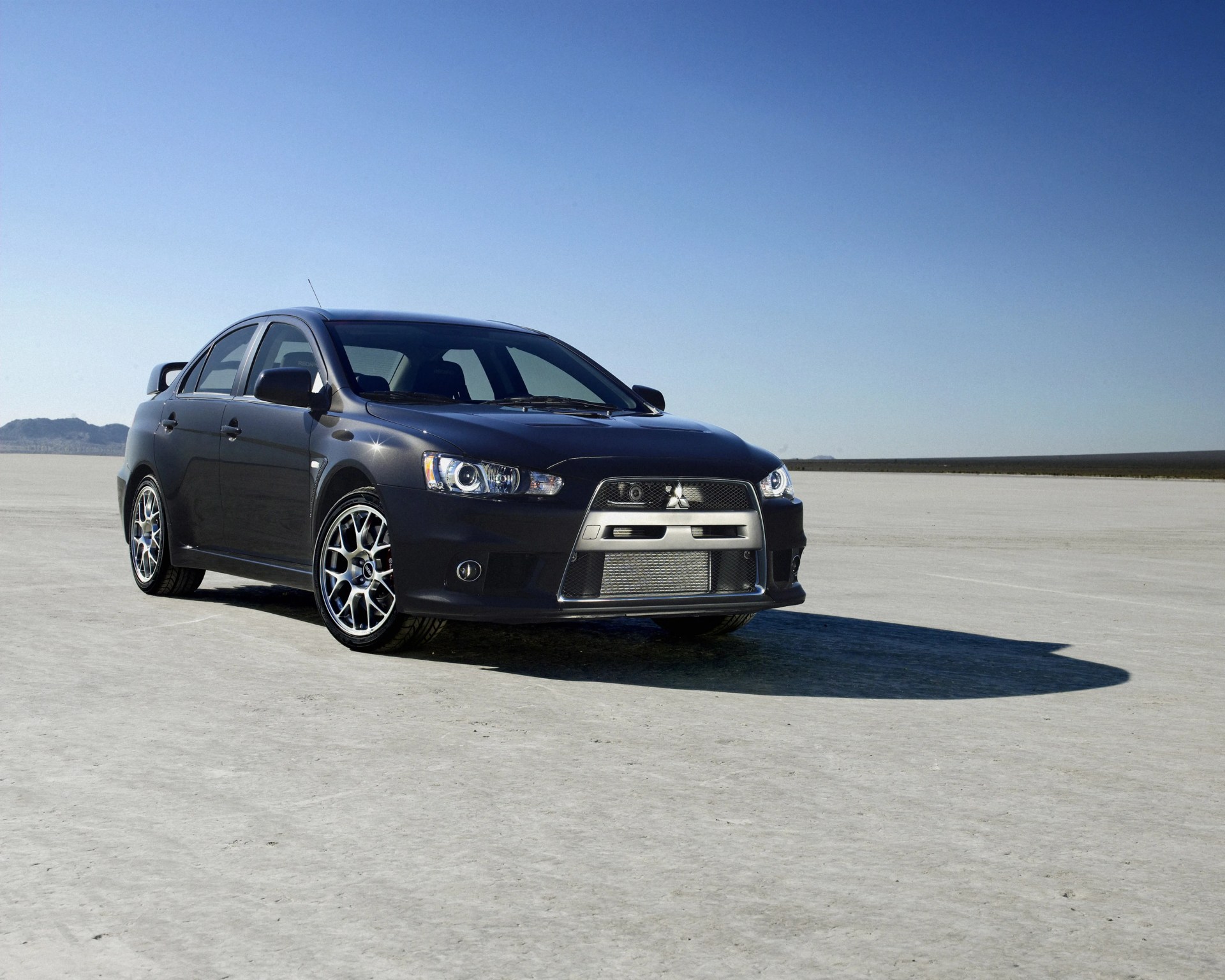 Black Mitsubishi Lancer Evolution sedan parked on a flat, dry desert surface under a clear blue sky.