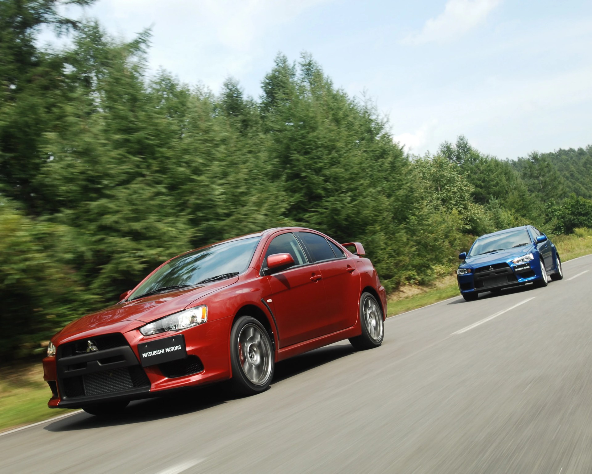 Red and blue Mitsubishi Lancer Evolution cars driving on a road with green trees in the background.