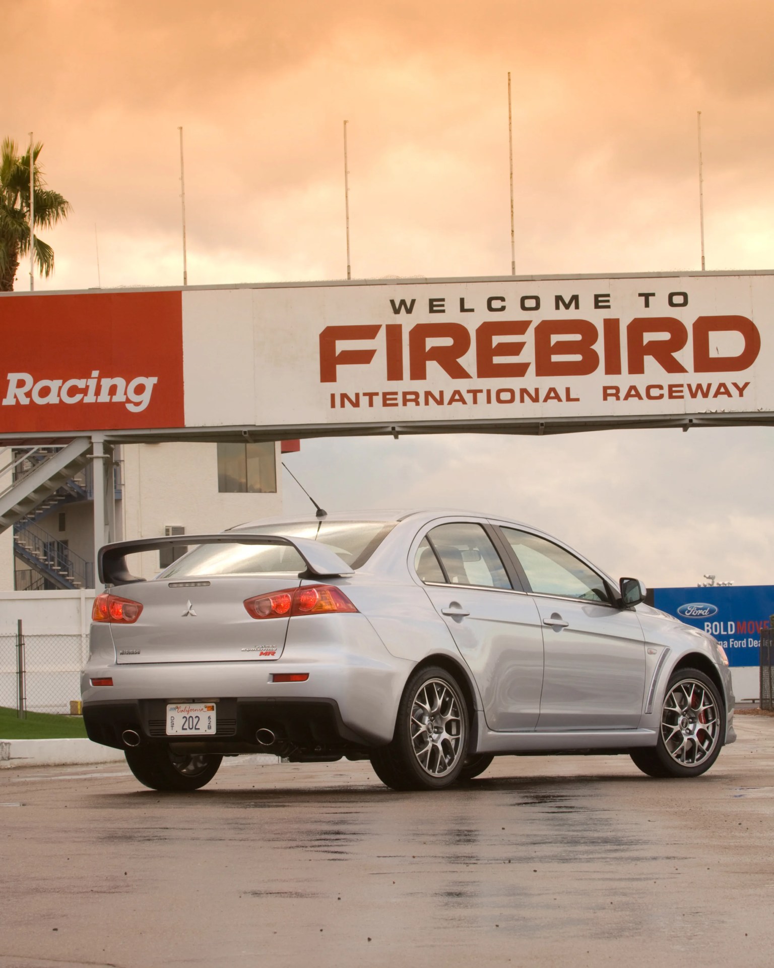 Silver Mitsubishi Lancer Evolution MR sedan parked under a "Welcome to Firebird International Raceway" sign.