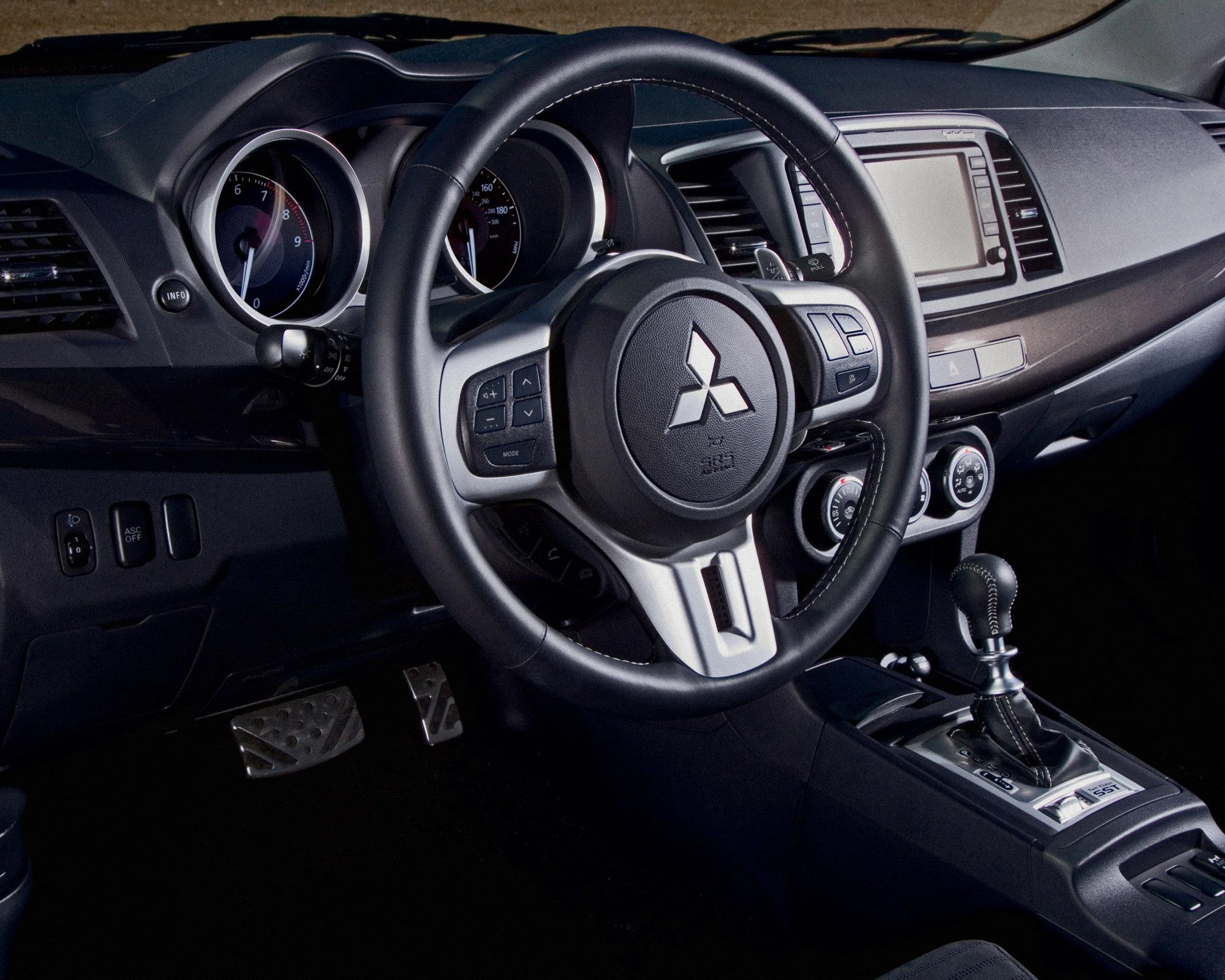 Black leather-wrapped Mitsubishi steering wheel with control buttons and dashboard featuring speedometer and infotainment screen.