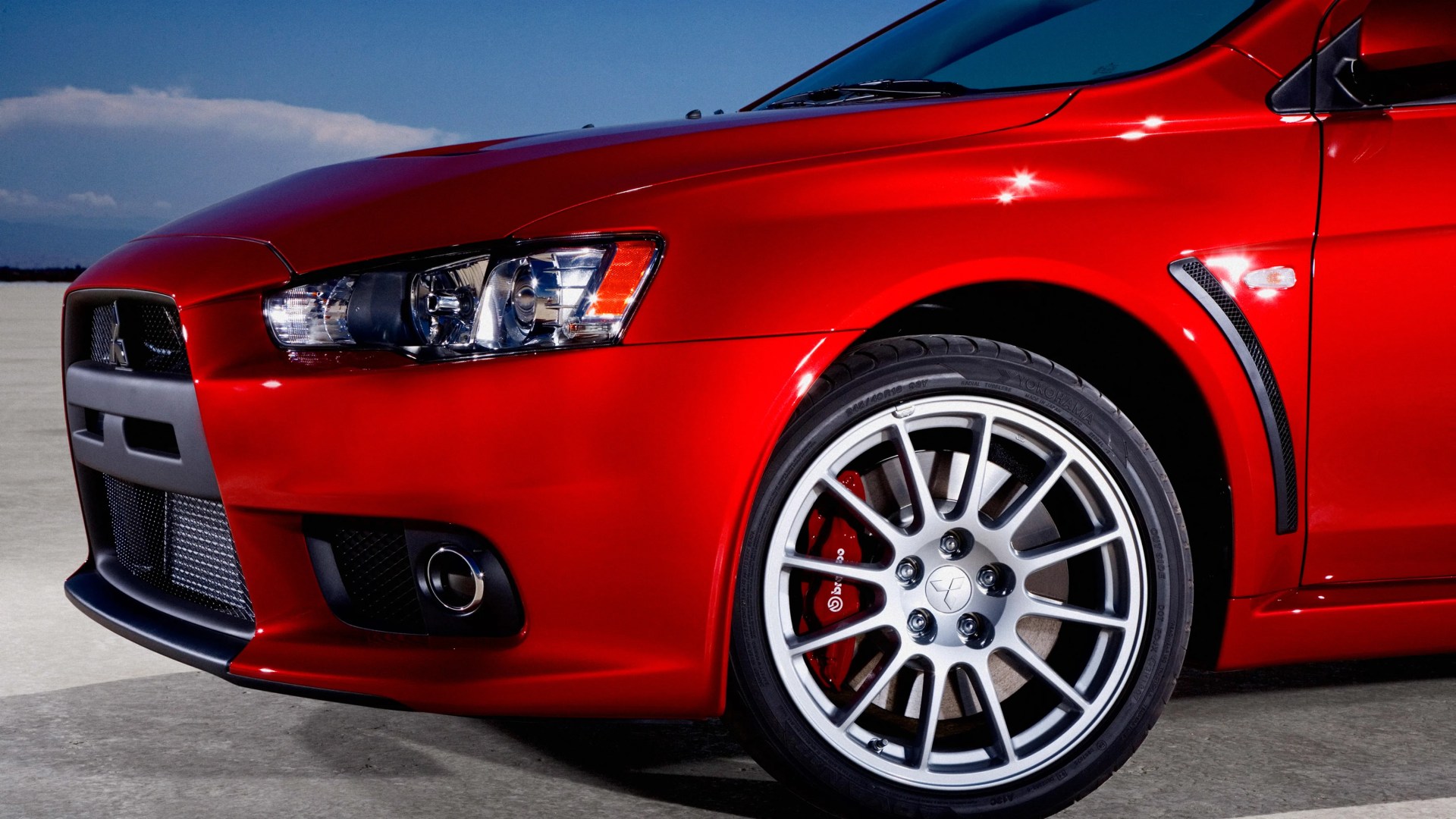 Front side view of a red Mitsubishi car showing the headlight, grille, and silver alloy wheel with Brembo brake caliper.