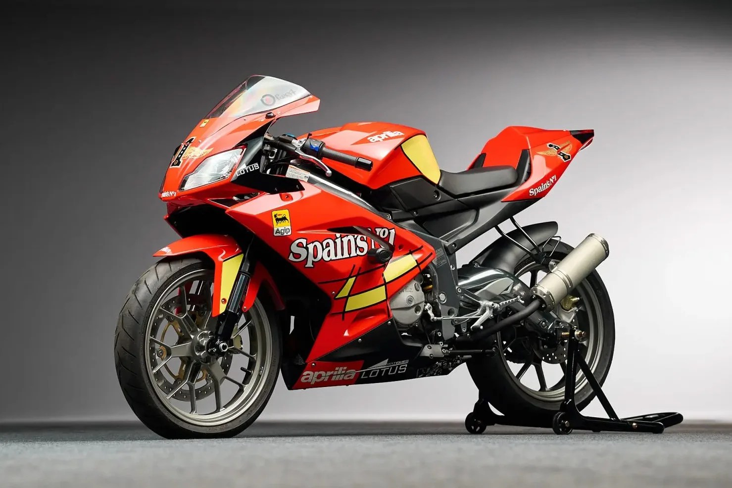 Red Aprilia racing motorcycle with Spain's No.1 decals and yellow accents on a gray studio floor.