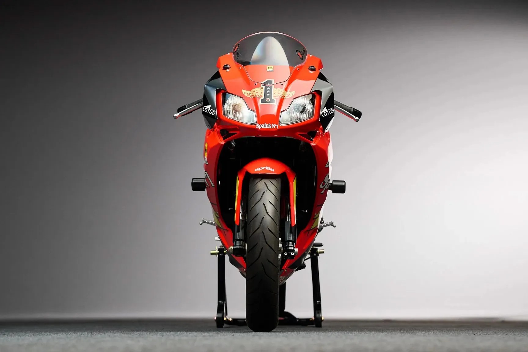 Front view of a red Aprilia racing motorcycle with black and yellow accents on a gray background.