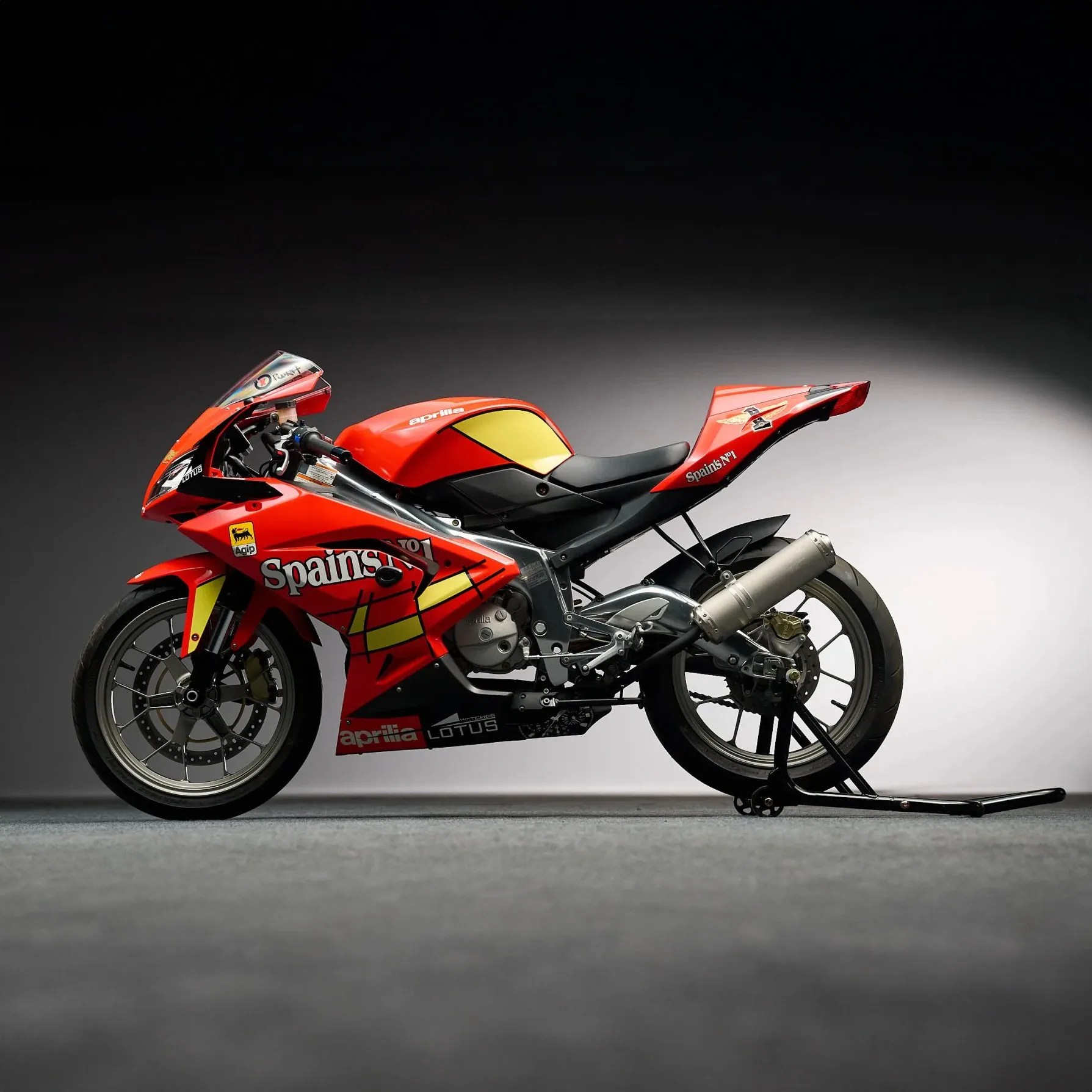Red Aprilia racing motorcycle with yellow and black accents on a gray floor against a dark background.
