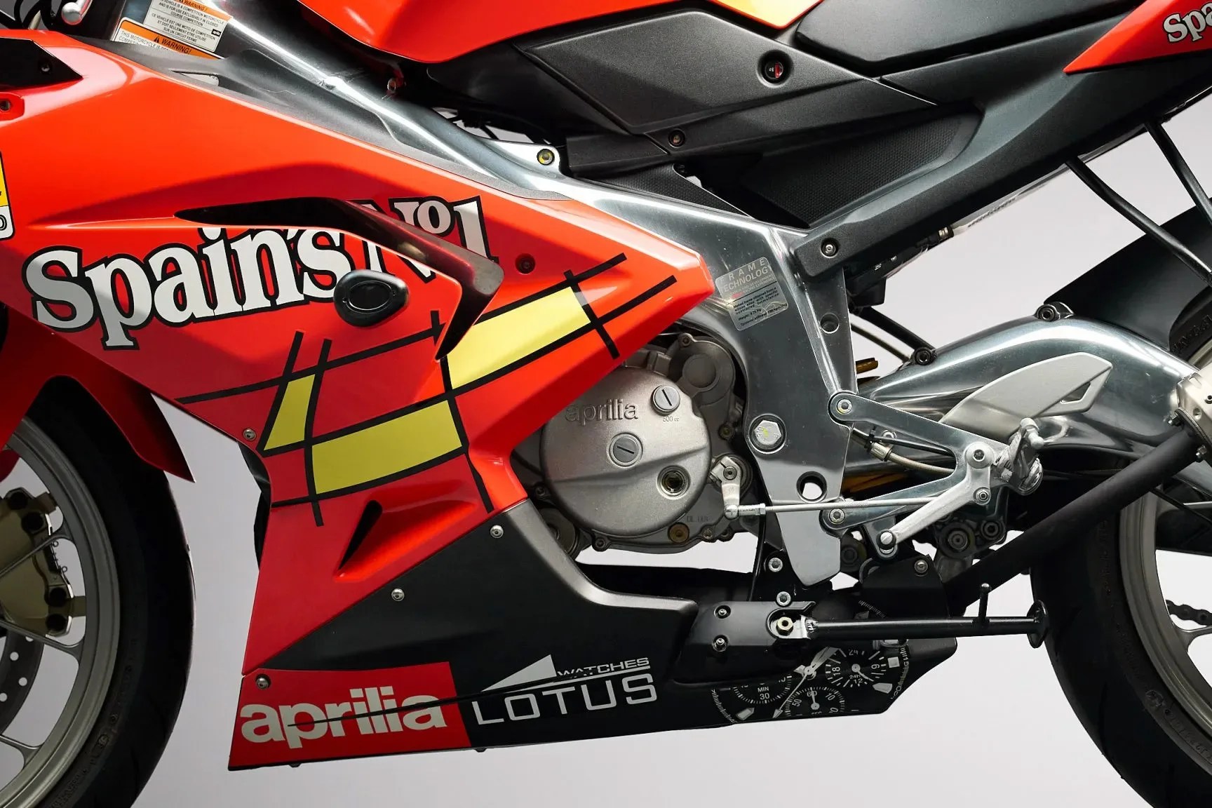 Close-up of the red Aprilia racing motorcycle fairing with Spain's No.1 and Lotus logos, showing engine and frame details.