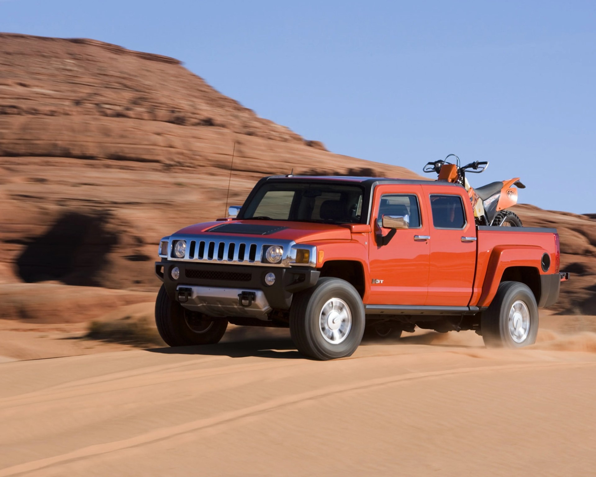 Orange Hummer H3T pickup truck driving on desert sand with an orange dirt bike in the bed.