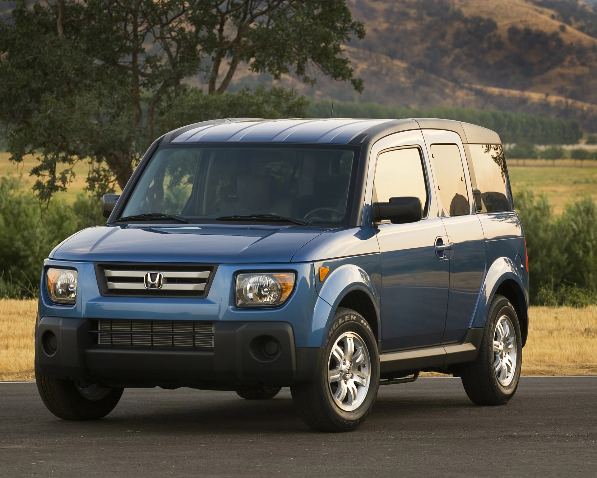 Blue Honda Element SUV with black trim parked on a road with trees and hills in the background.