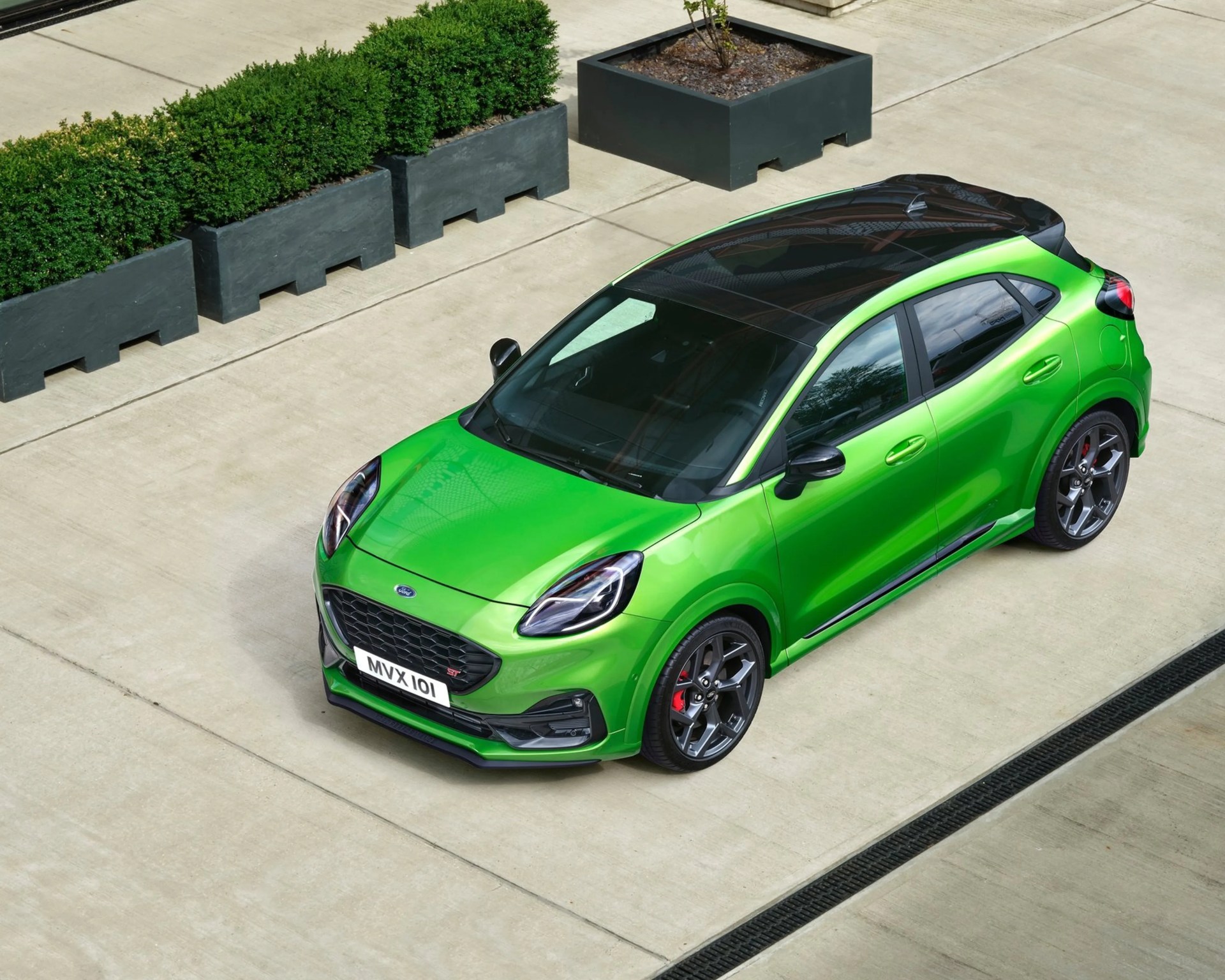 Bright green Ford Puma ST with black roof and dark alloy wheels parked on concrete near large black planters with green shrubs.