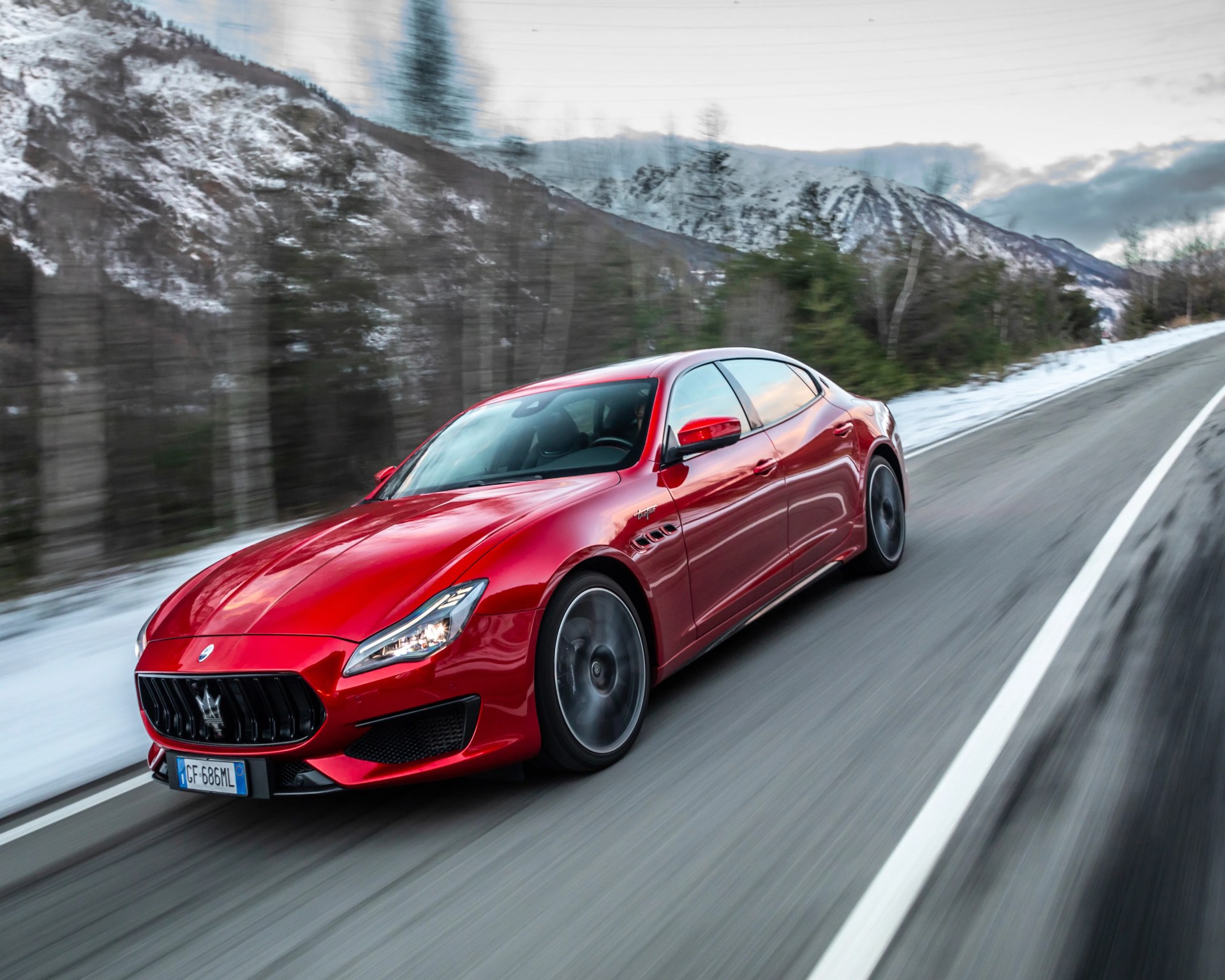 Red Maserati sports car driving on a snowy mountain road with blurred trees and mountains in the background.