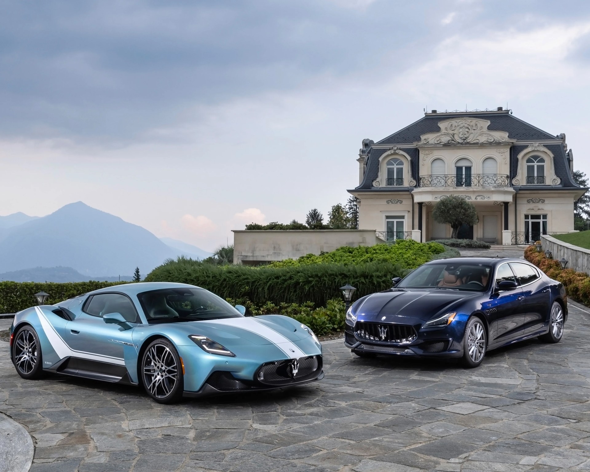 Two Maserati cars, a light blue sports car and a dark blue sedan, parked on a stone driveway in front of a large ornate house.