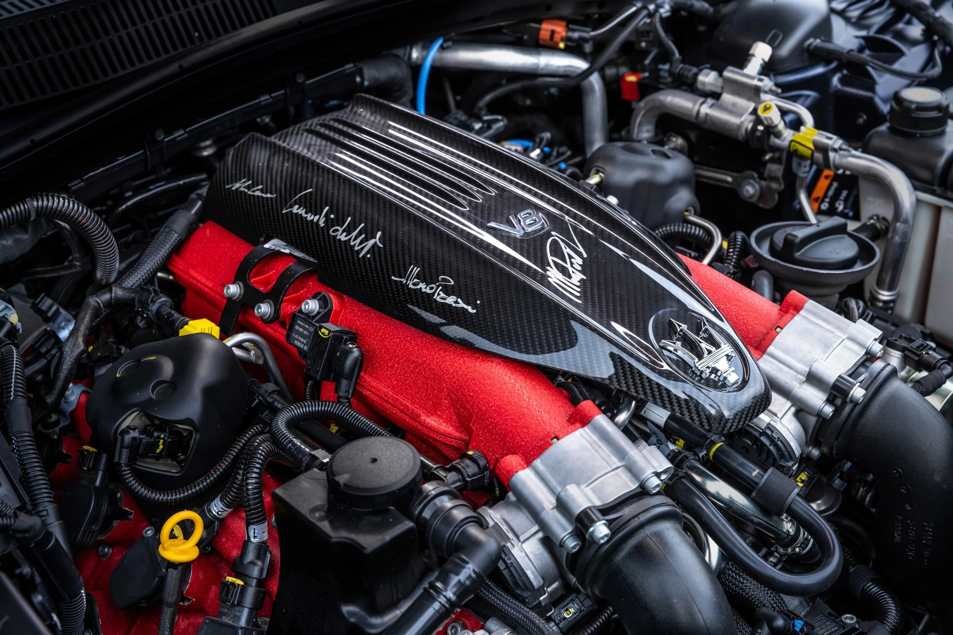 Carbon fiber engine cover with V8 emblem and Maserati trident logo on red engine components.