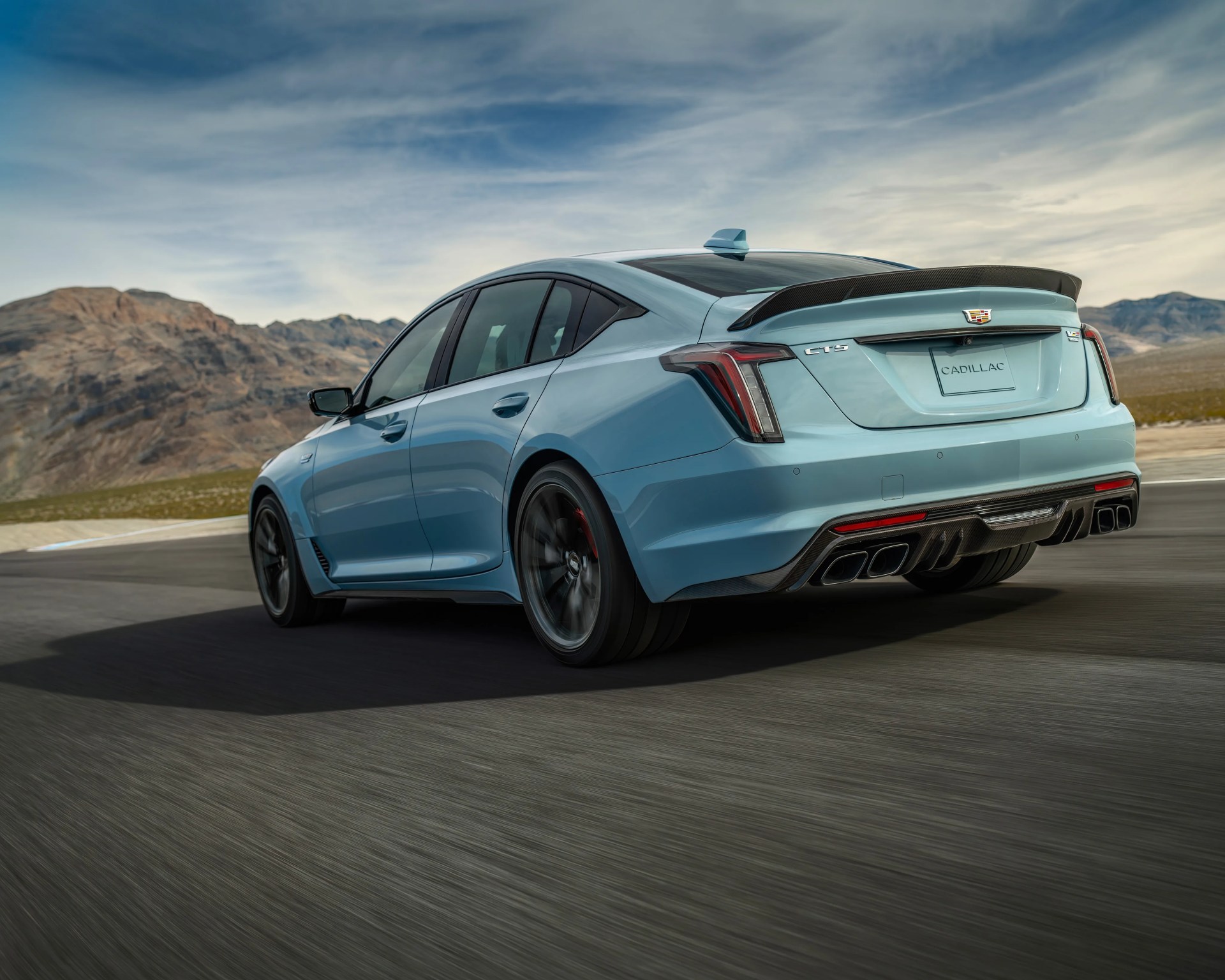Light blue Cadillac CT5 V-Series sedan with black rear spoiler and quad exhausts on a road with mountains in the background.