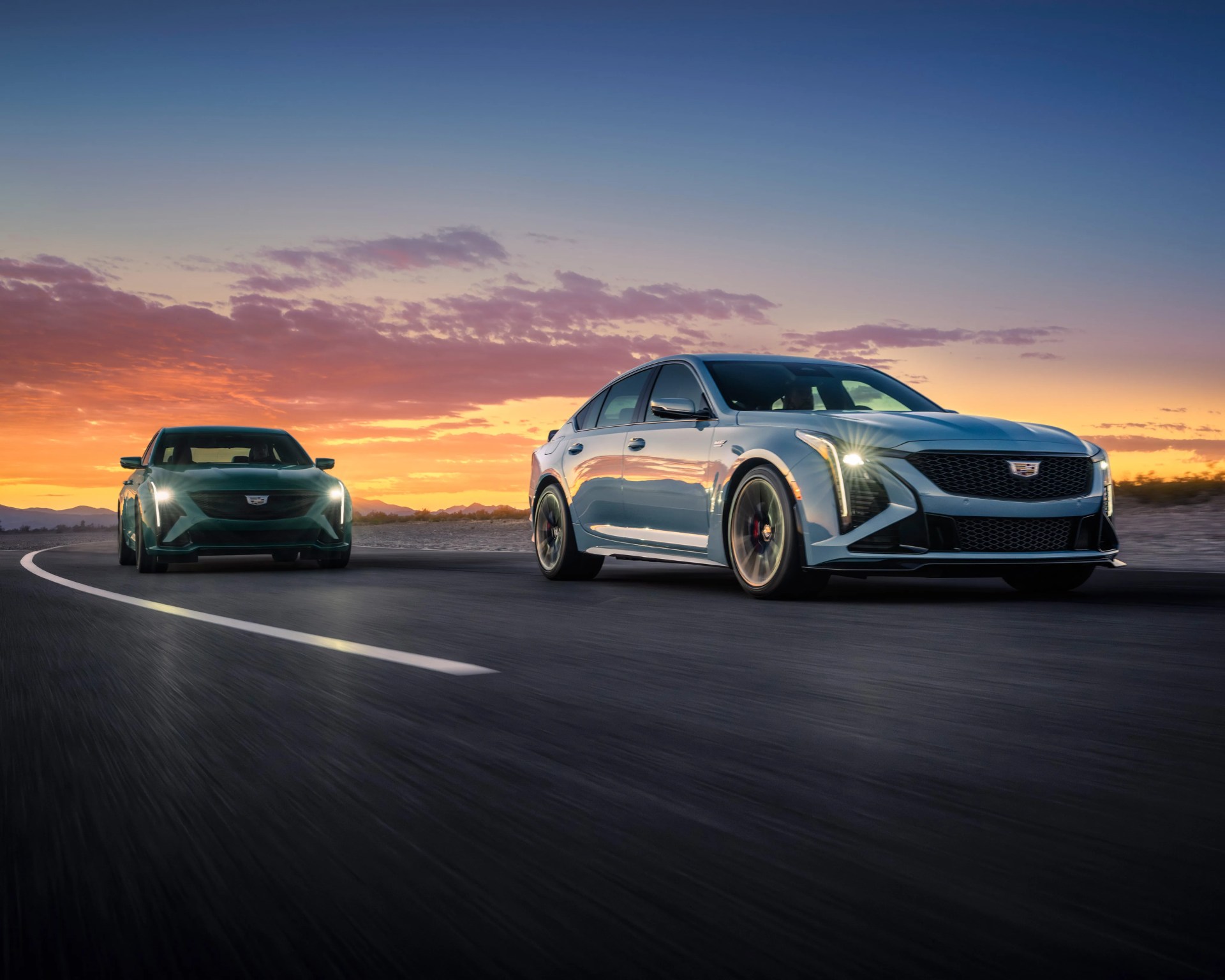 Two Cadillac sedans with headlights on driving on a curved road at sunset.