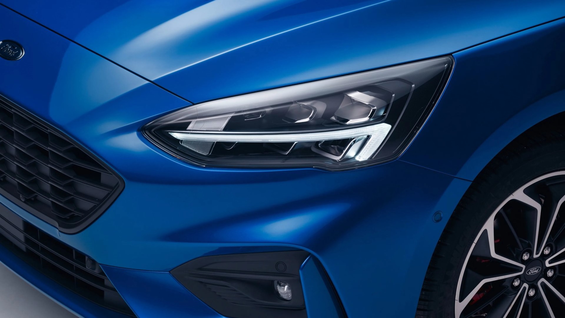 Close-up of a blue Ford car's front headlight, grille, and part of a black and silver alloy wheel.