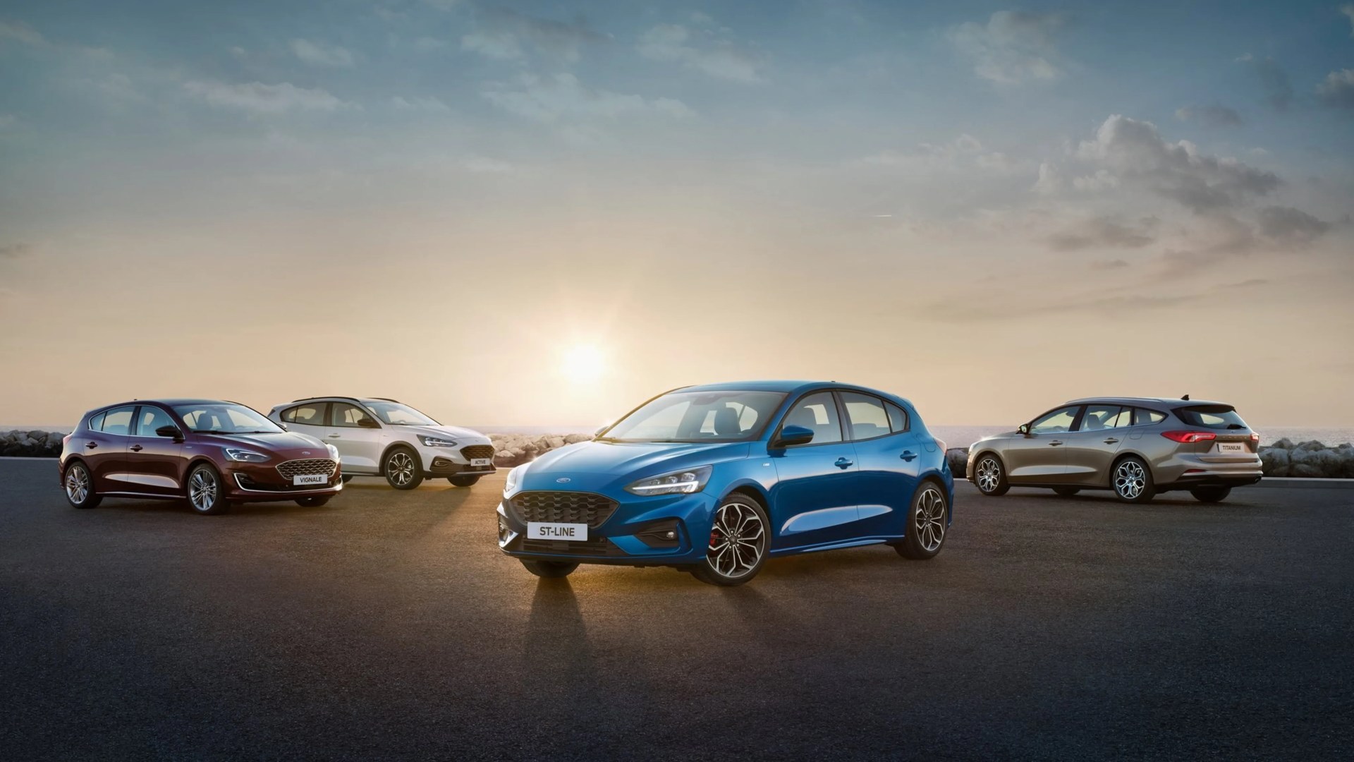 Four Ford hatchback cars in blue, maroon, white, and beige parked on a paved surface at sunset.
