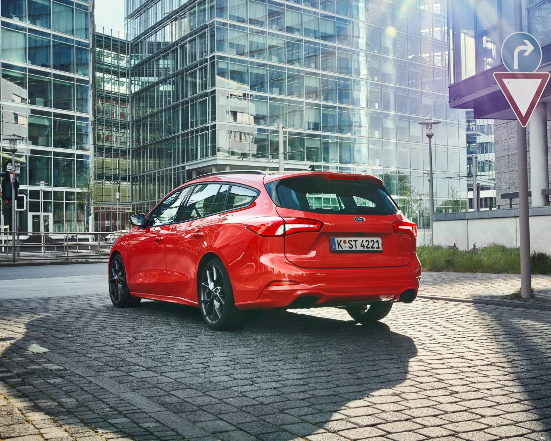 Red Ford station wagon parked on a cobblestone street in front of glass office buildings.
