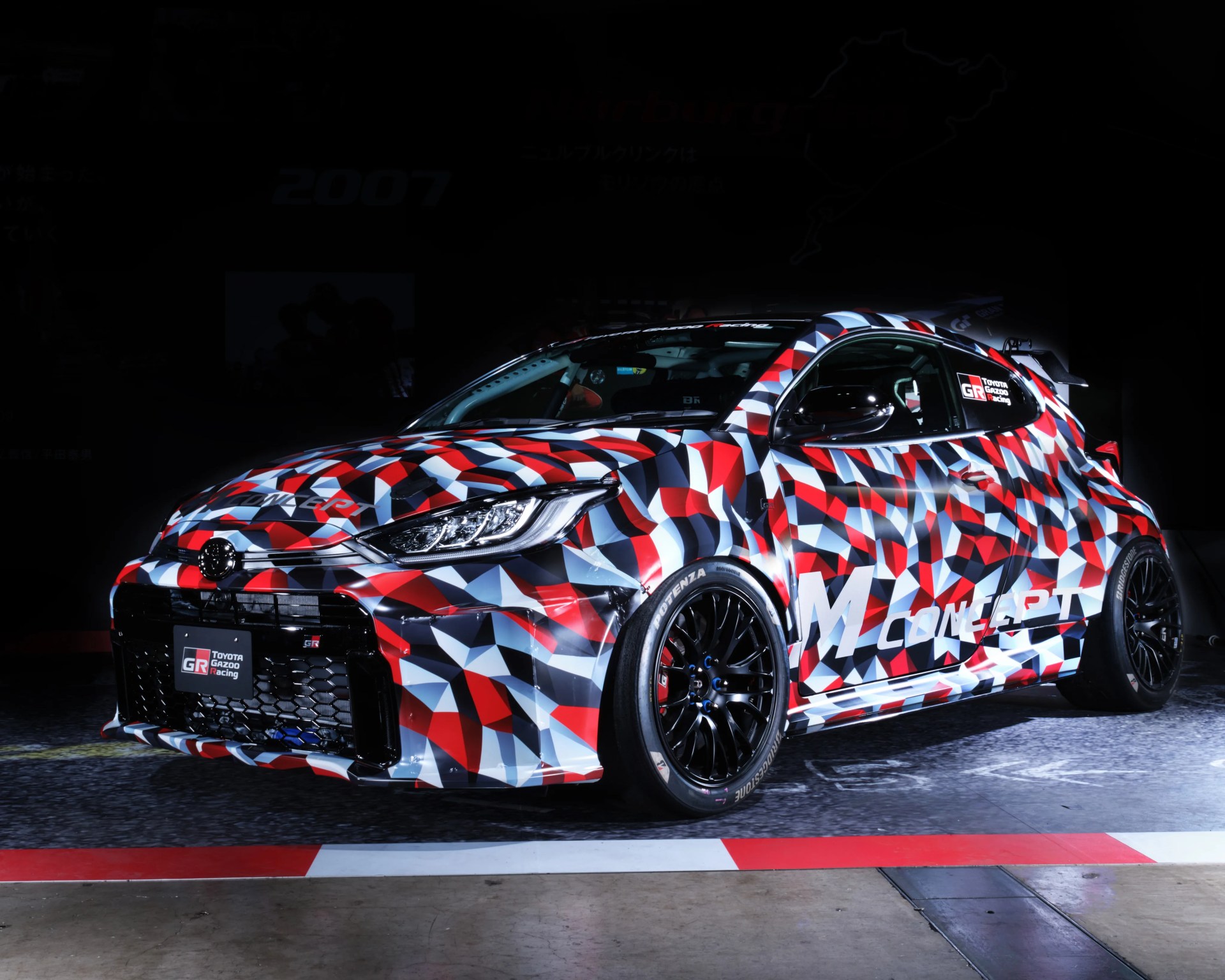 Toyota GR racing car with red, white, black, and blue geometric camouflage pattern on a racetrack.