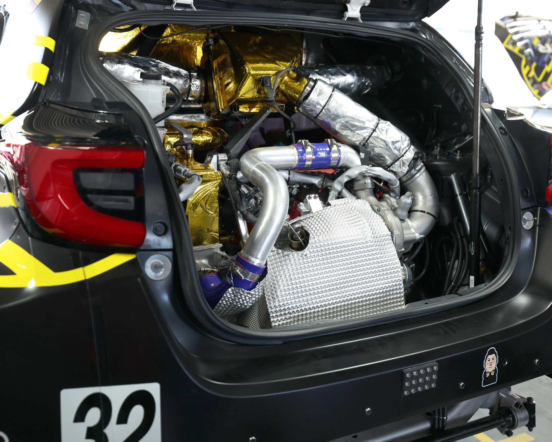 Rear view of a race car engine bay with metallic silver and gold heat shielding and blue silicone hose connectors.