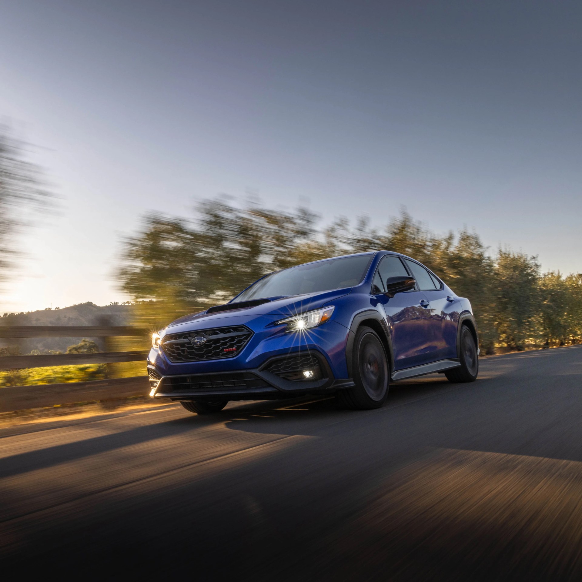 Blue Subaru WRX driving on a road with blurred trees and hills in the background at sunset.