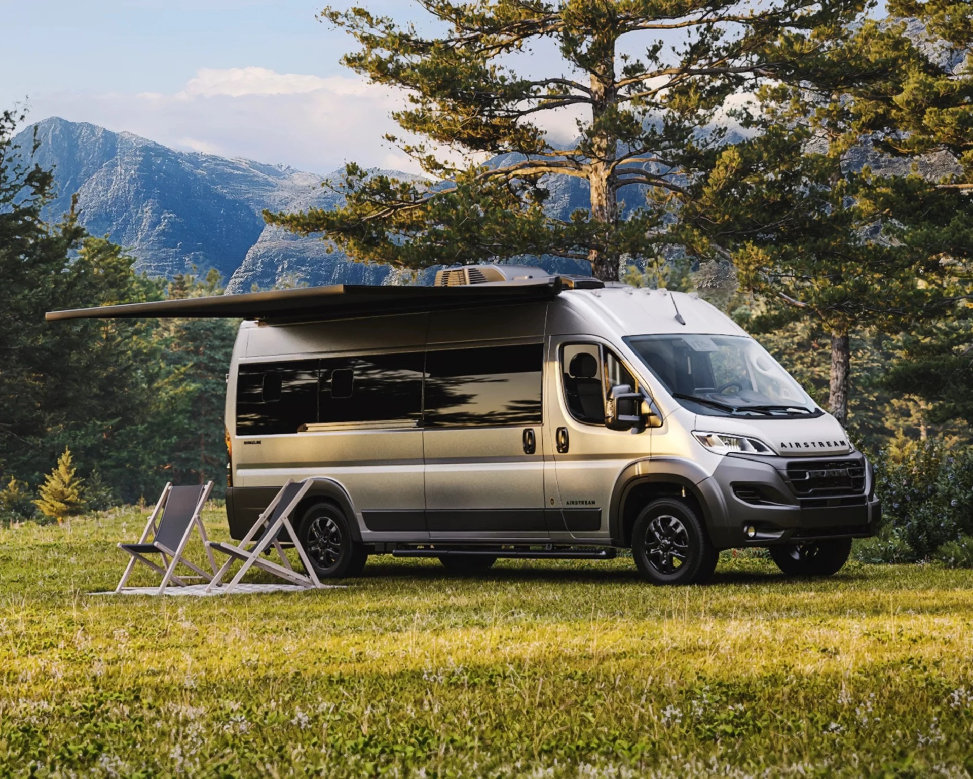 Silver Airstream camper van with extended awning parked on grass near two folding chairs and pine trees.