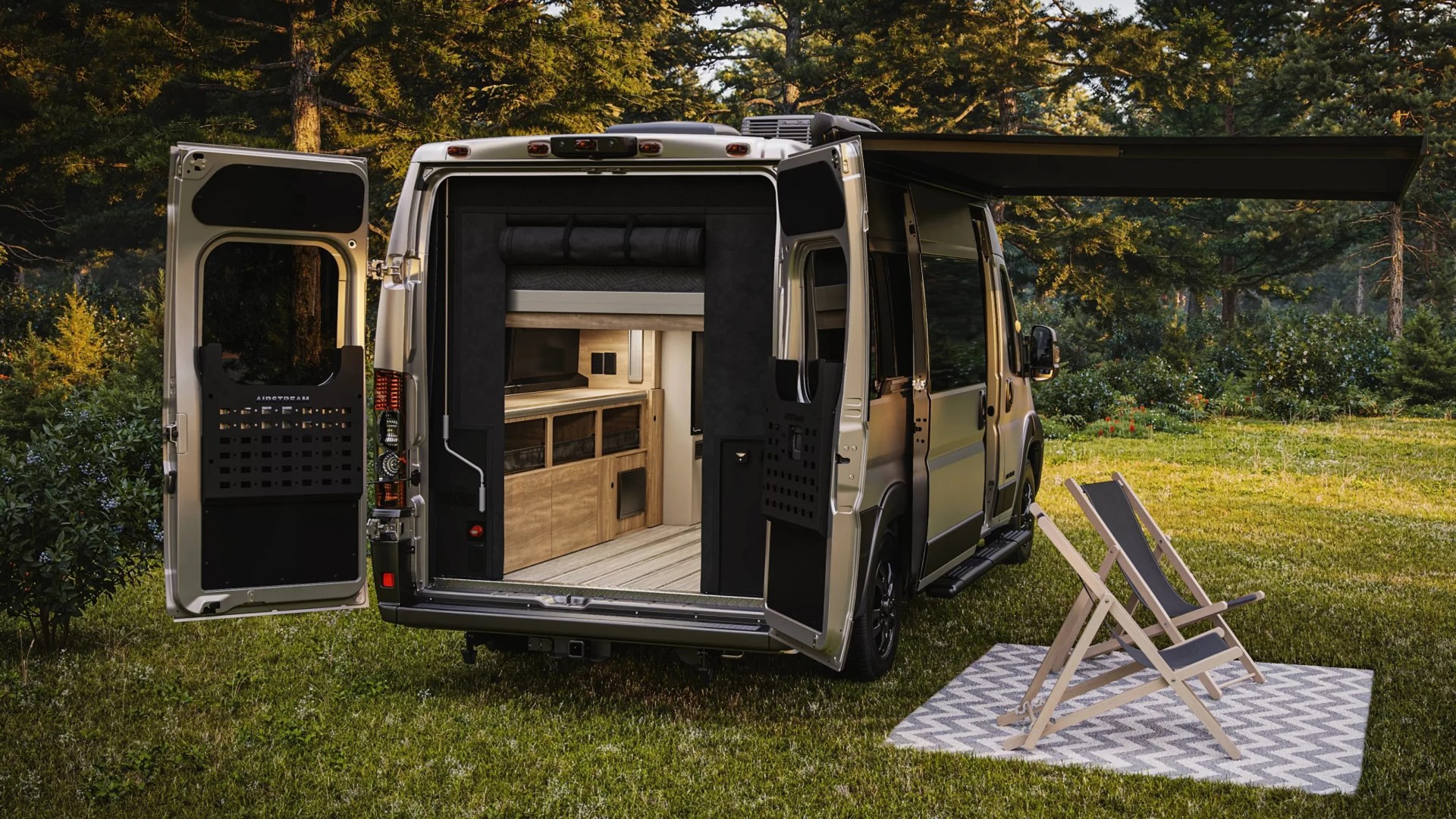 Silver camper van with rear doors and side awning open, showing wooden interior, parked on grass near two folding chairs on a patterned rug.