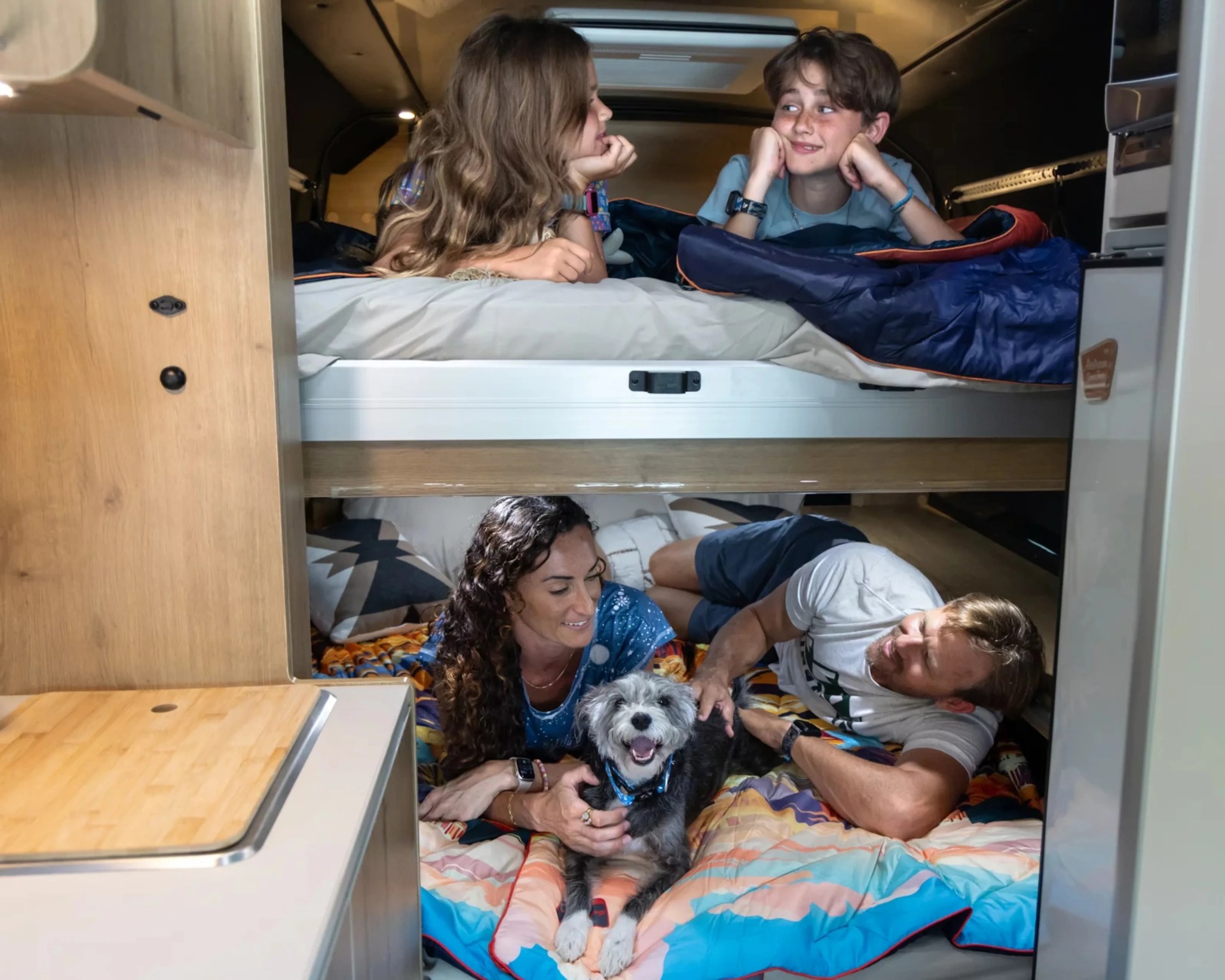 Two children lying on an upper bunk bed and two adults with a dog on a lower bunk bed inside a camper van.